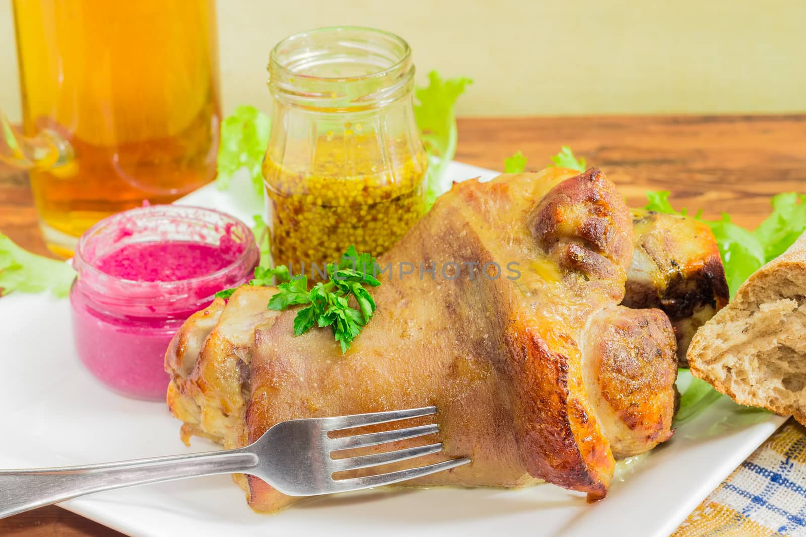 Baked ham hock with fork on a white dish on the background of a beer and condiments closeup
