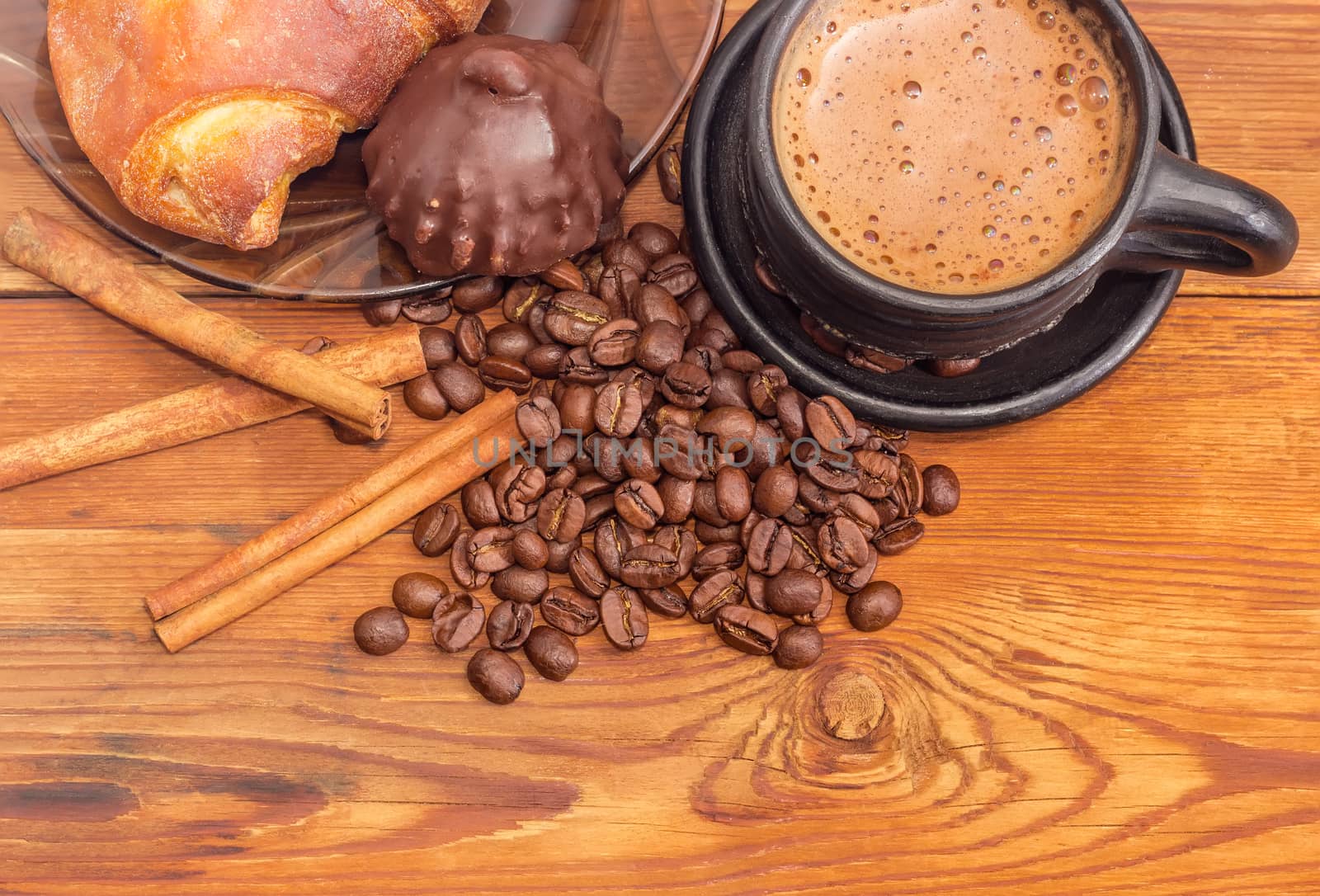 Fragment of black ceramic cup with freshly brewed coffee closeup on the background of roasted coffee beans, cinnamon sticks, chocolate truffle and croissant on a wooden surface
