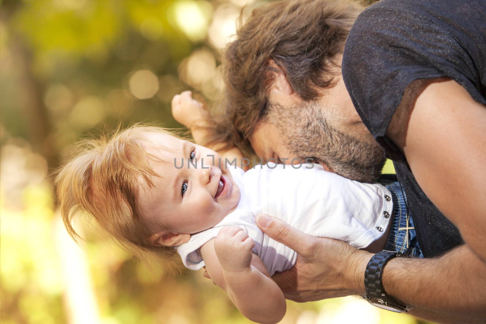 A cute little baby is laughing while his father is biting him playfully outdoors.