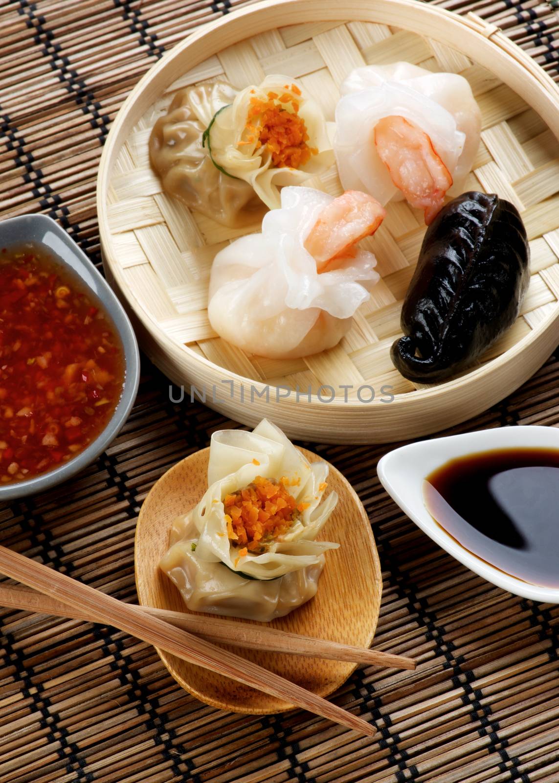 Assorted Dim Sum in Bamboo Steamed Bowl and Tori with Chiken on Wooden Plate with Red Chili and Soy Sauces and Chopsticks closeup on Straw Mat background