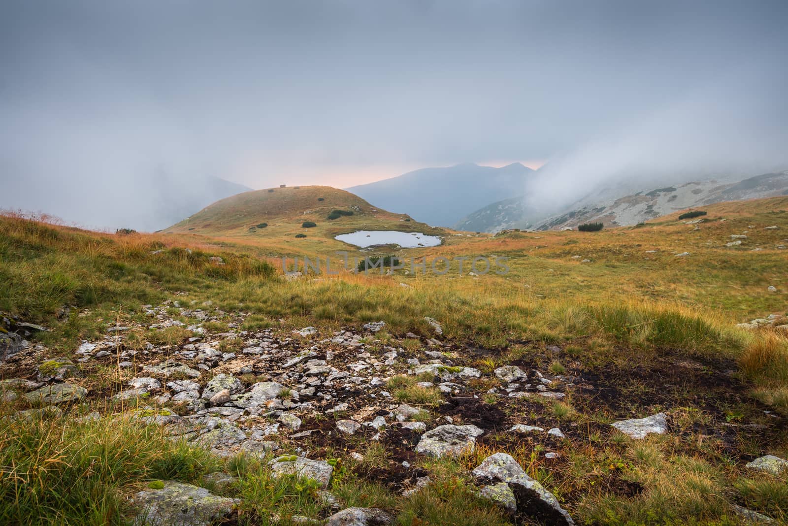 Foggy Mountain Landscape with a Tarn and Rocks in Foreground at Sunset