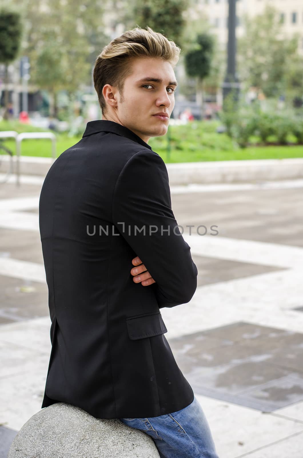 Serious or sad blond young man in jeans and jacket, sitting outdoors looking in camera