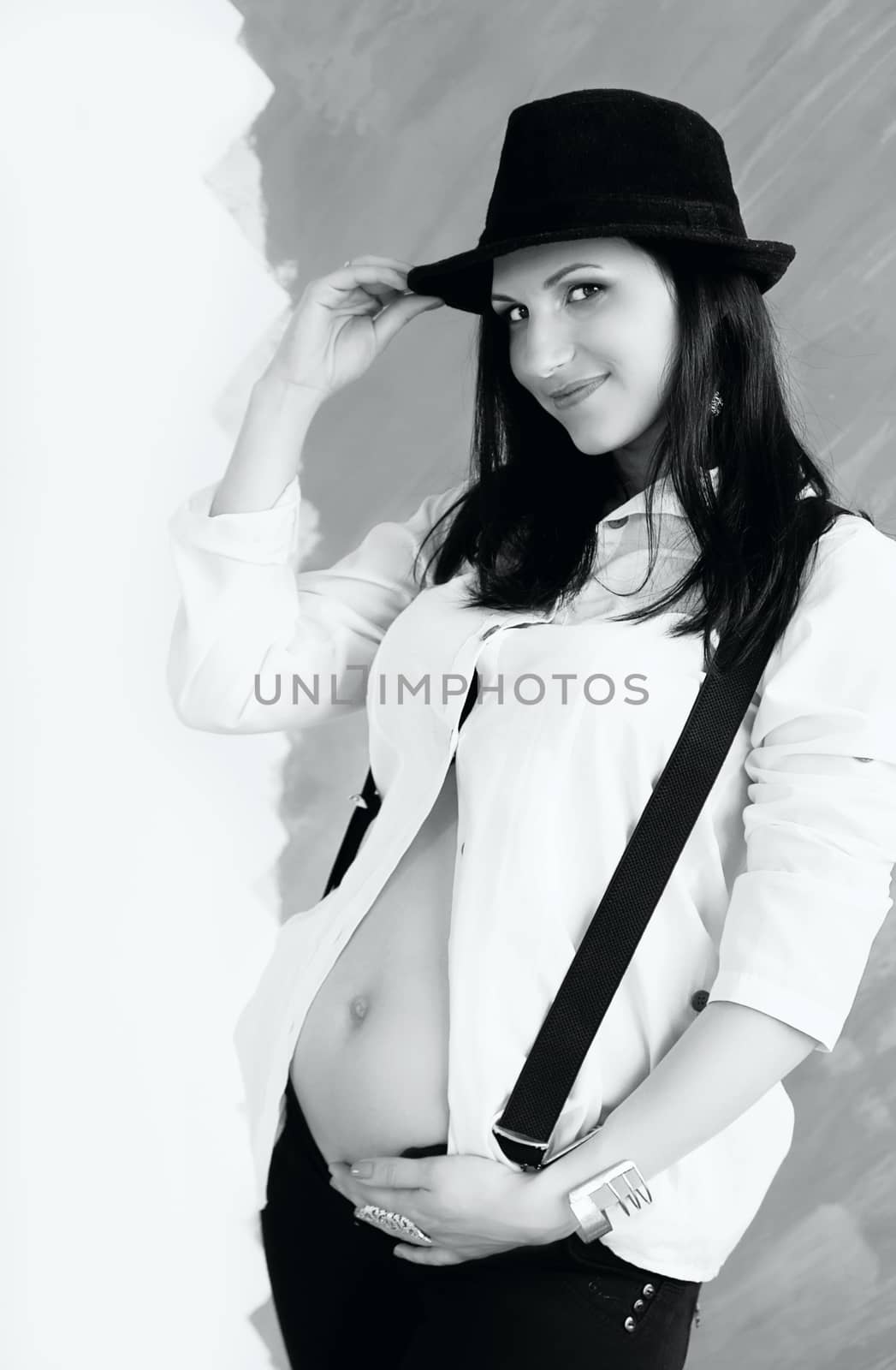 Portrait of the young pregnant woman in white shirt with suspenders and hat. Black and white photography