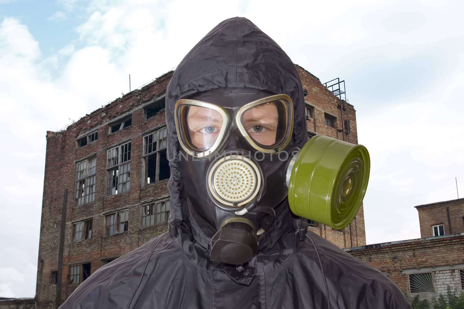 Head and shoulders of a person in a rubber gas mask and in a black jacket with a hood against the backdrop of abandoned and partially destroyed industrial building
