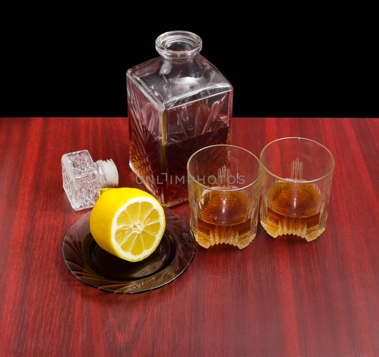 Decanter and two glasses with whiskey, stopper from the decanter and lemon on a saucer on a wooden table on a dark background
