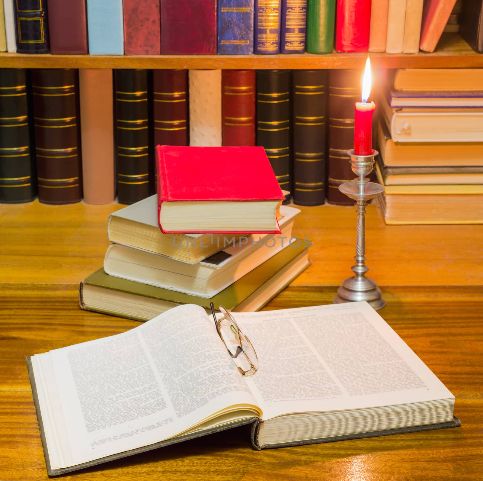 Open big book and glasses, stack of books different formats and cover design on a wooden table against the background of shelf with books by candlelight
