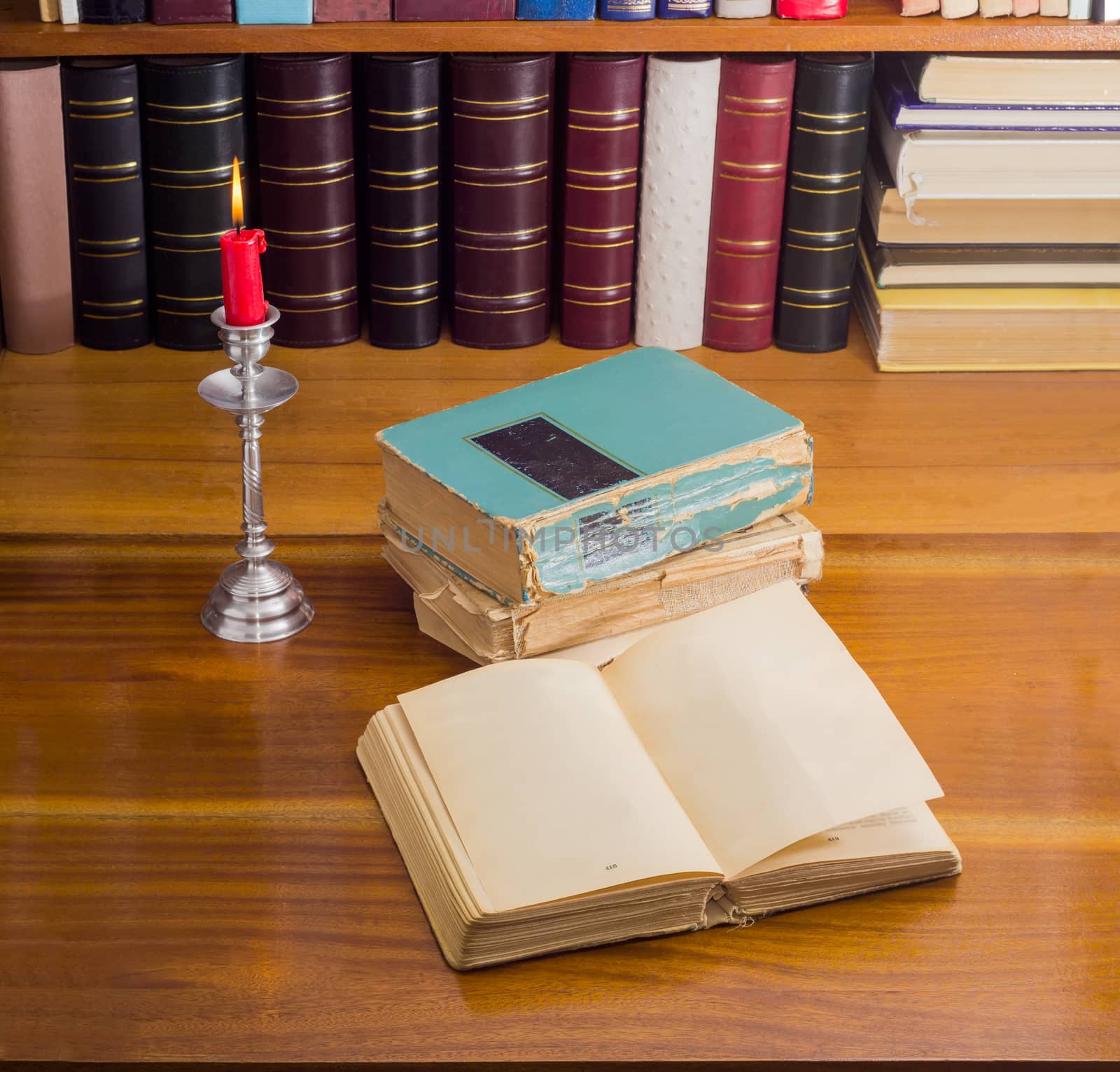 Open old book, stack of other old shabby books and a burning candle in candlestick on a wooden table against the background of shelf with books
