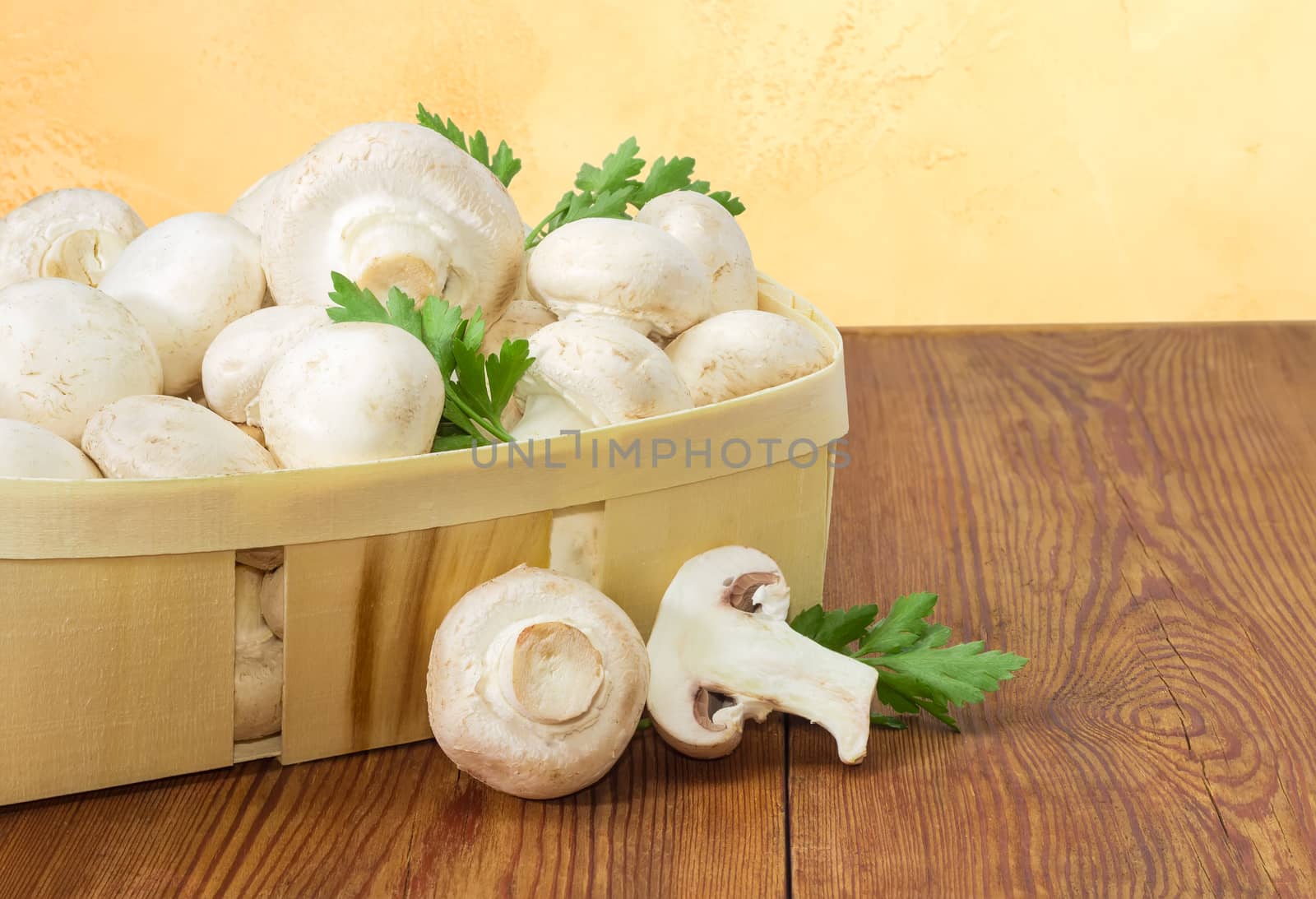 Fragment of wooden basket with fresh cultivated button mushrooms and twigs of parsley, one whole mushroom and one half of mushroom separately beside on an old wooden surface
