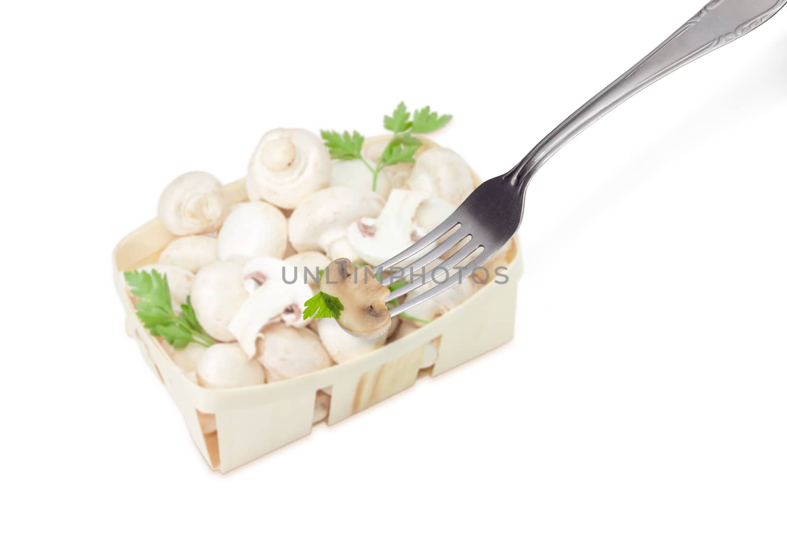 Half of the fried button mushroom with parsley leaf on the stainless steel fork on the background of blurred wooden basket with uncooked mushrooms on a light background
