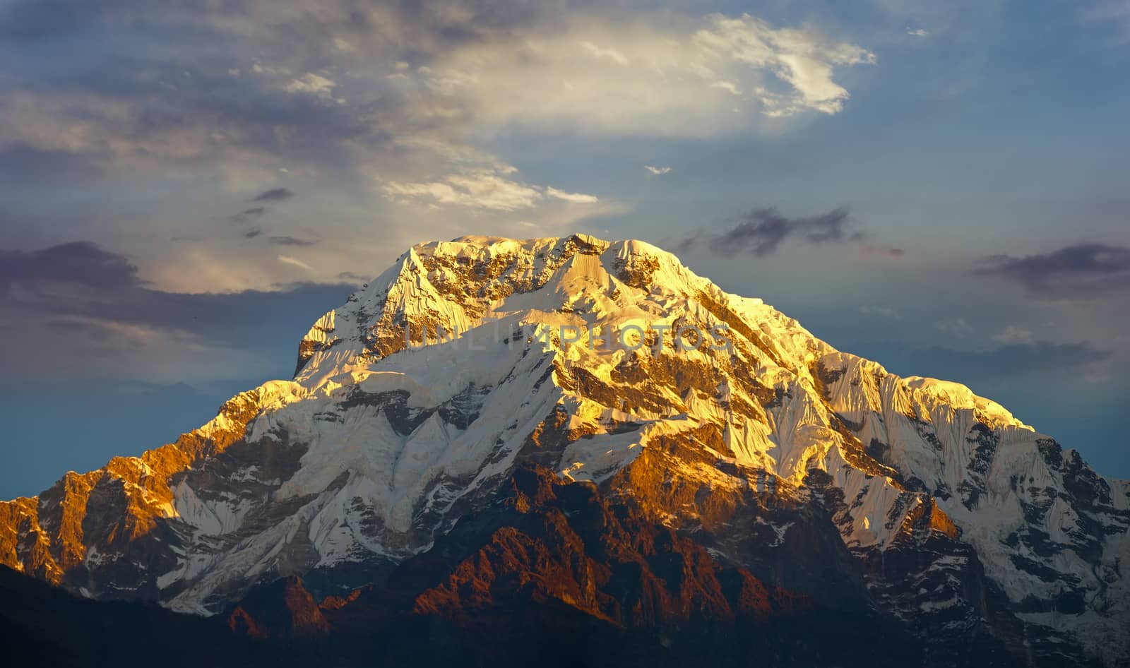 Mountain peak covered with glaciers in the rays of the rising sun against the backdrop of sky with clouds in the Himalayas
