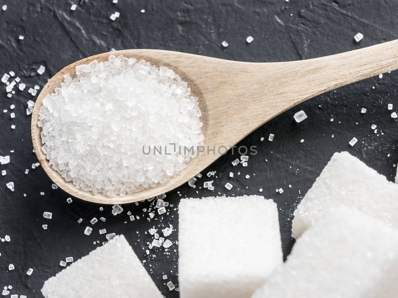 Granulated sugar in wooden spoon close up. Background of sugar cubes and sugar in spoon. White sugar on black background.
