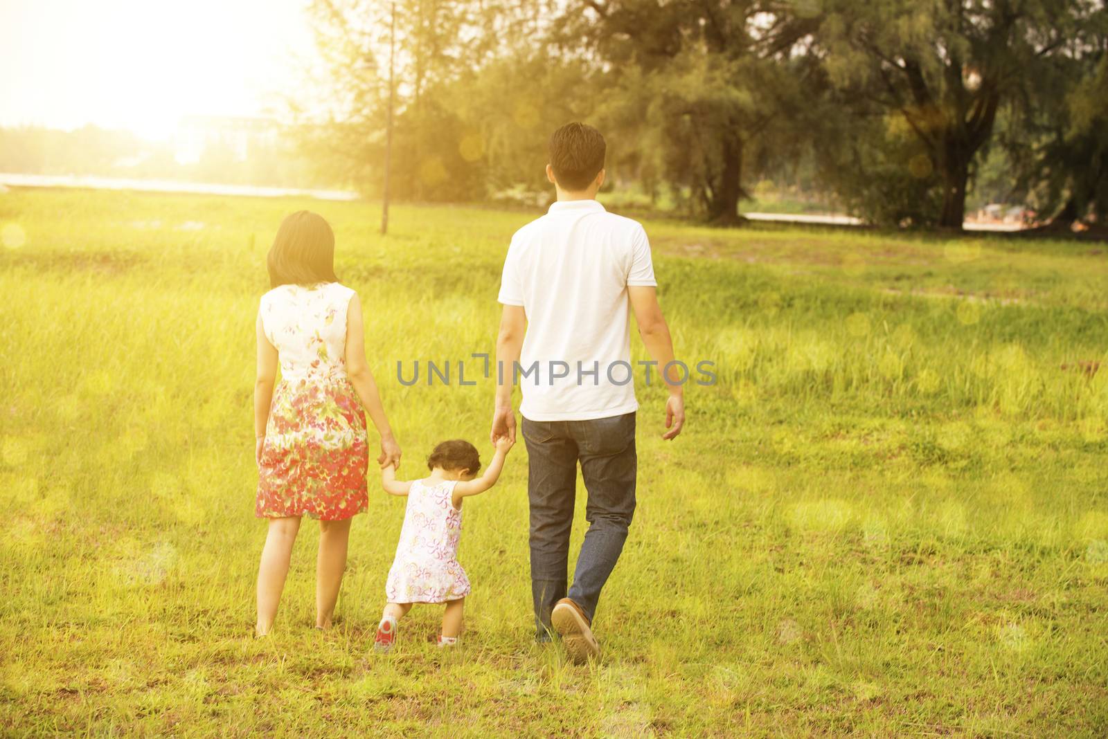 Rear view of Asian family enjoying outdoor activity together, walking on garden lawn in beautiful sunset during holiday vacations.