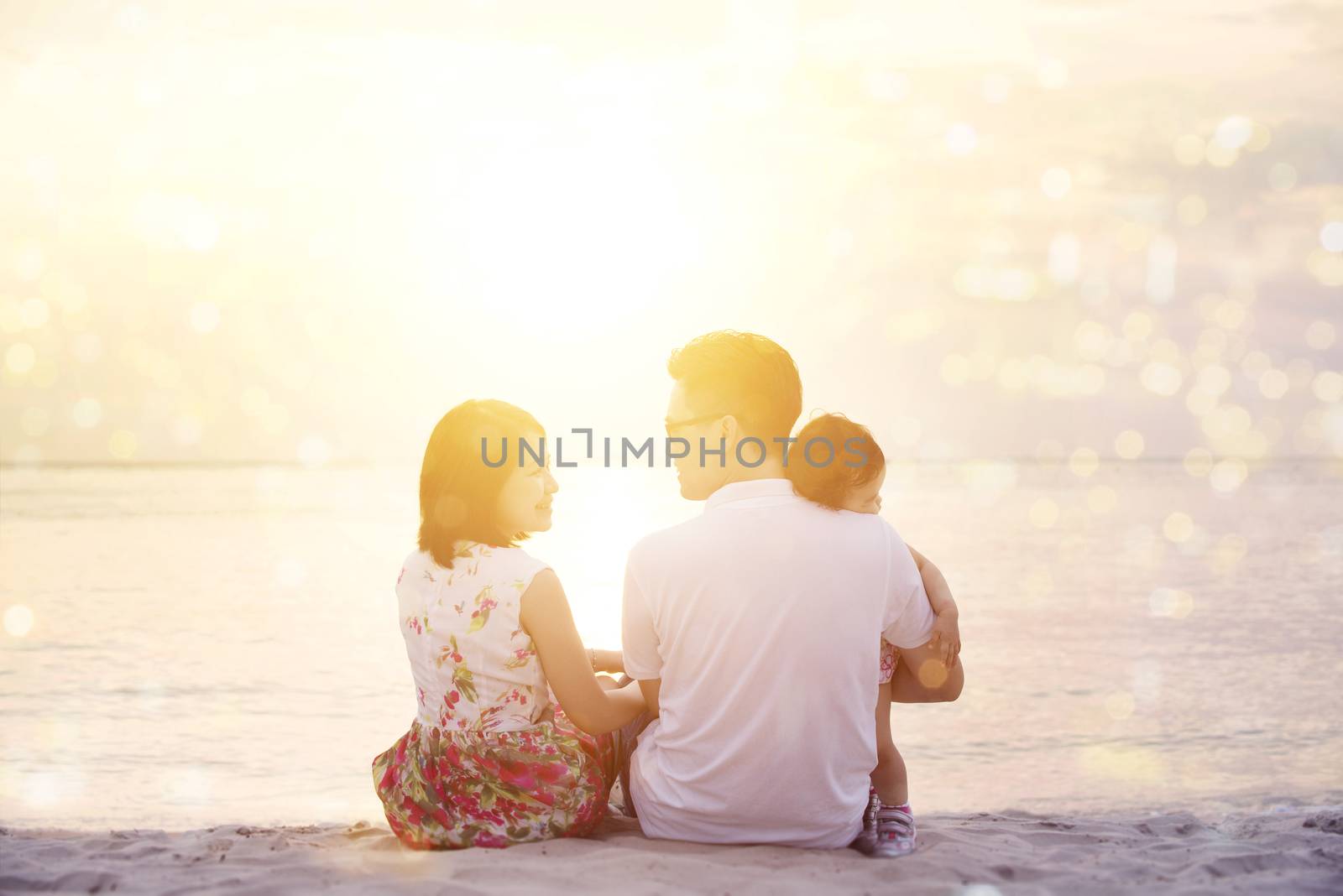 Rear view of family enjoying outdoor activity together, sitting on beach sand in beautiful sunset during holiday vacations.
