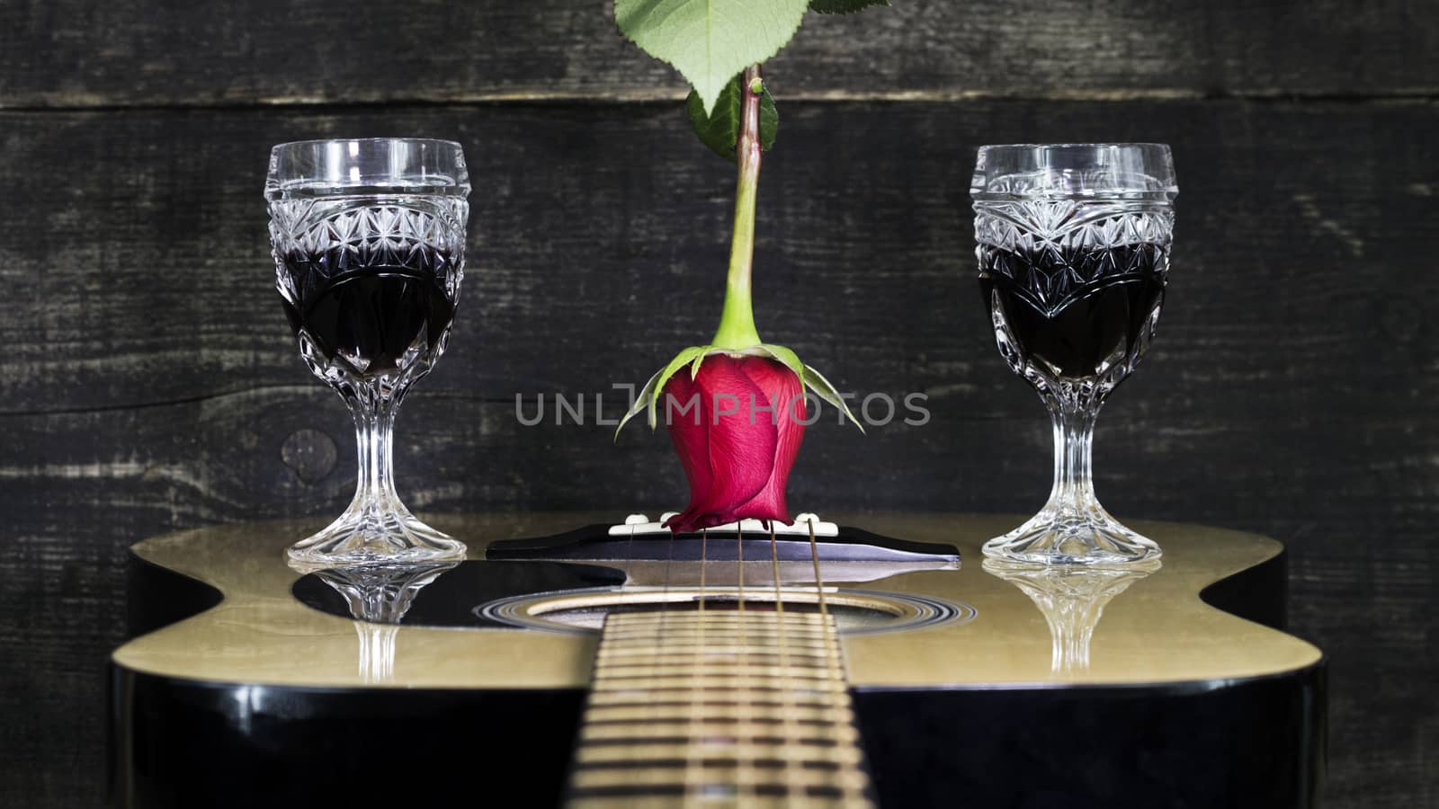 Red Rose and Wine Glasses Resting On Acoustic Guitar With Wooden Background