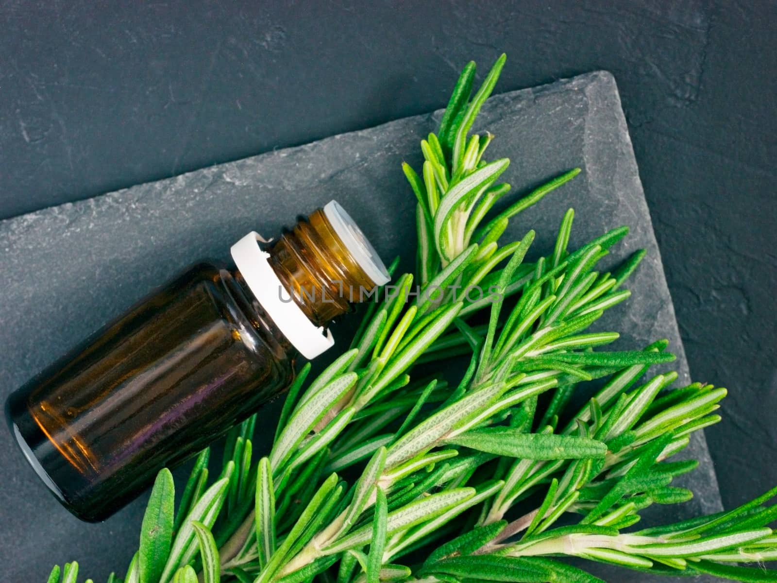 Rosemary essential oil in dark glass bottle and fresh rosemary on dark background. Top view or flat lay