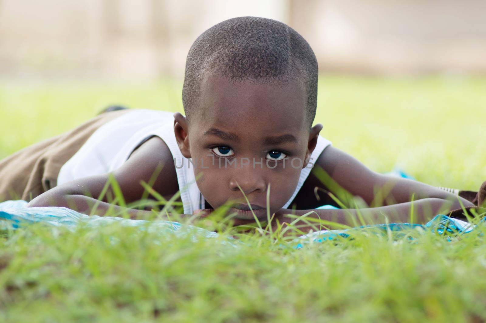 This child lying on a piece of cloth , looking at the camera , sits in the shade of a tree on the lawn.