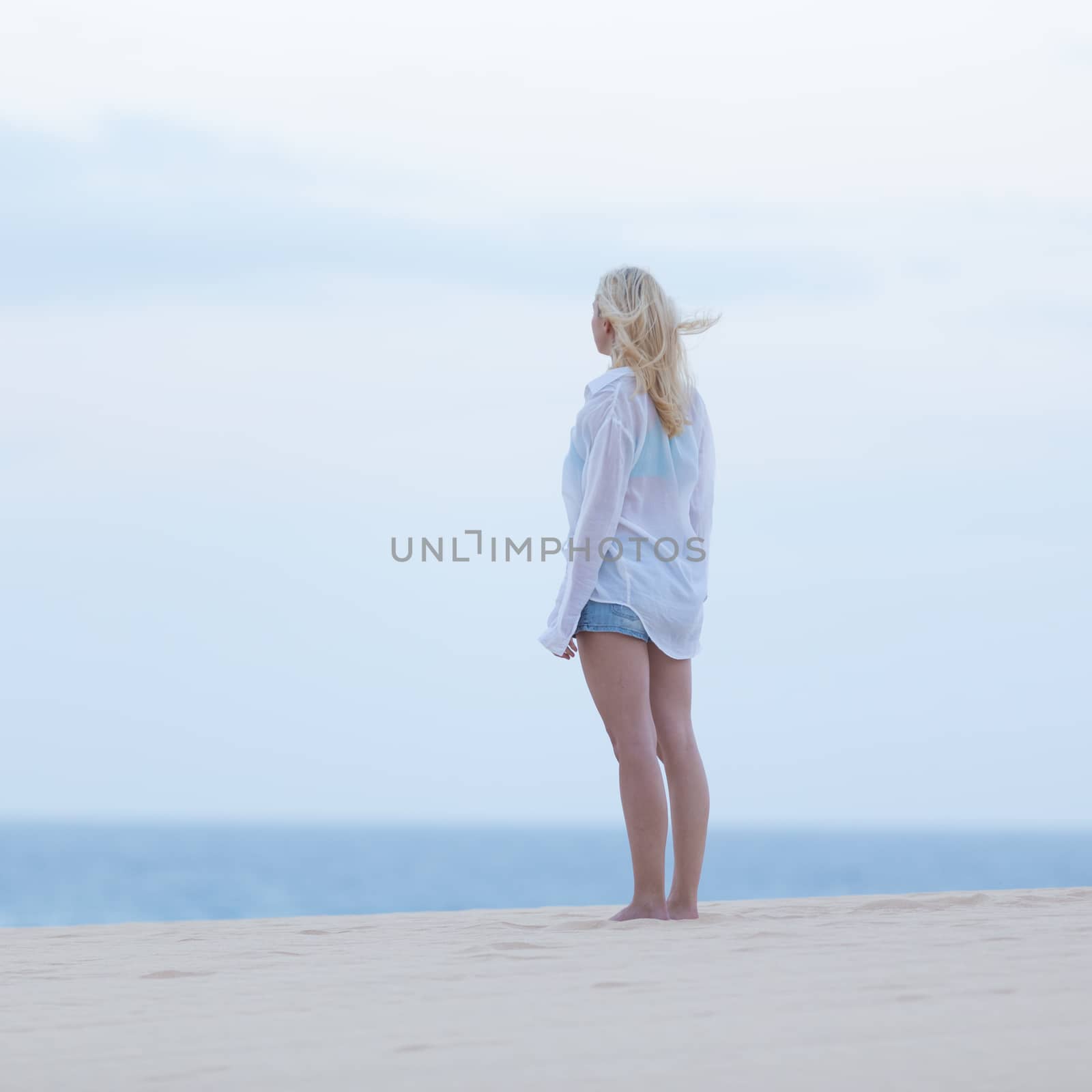 Meditative, sensual blonde woman wearing white loose casual shirt on vacations looking at horizon at dusk.