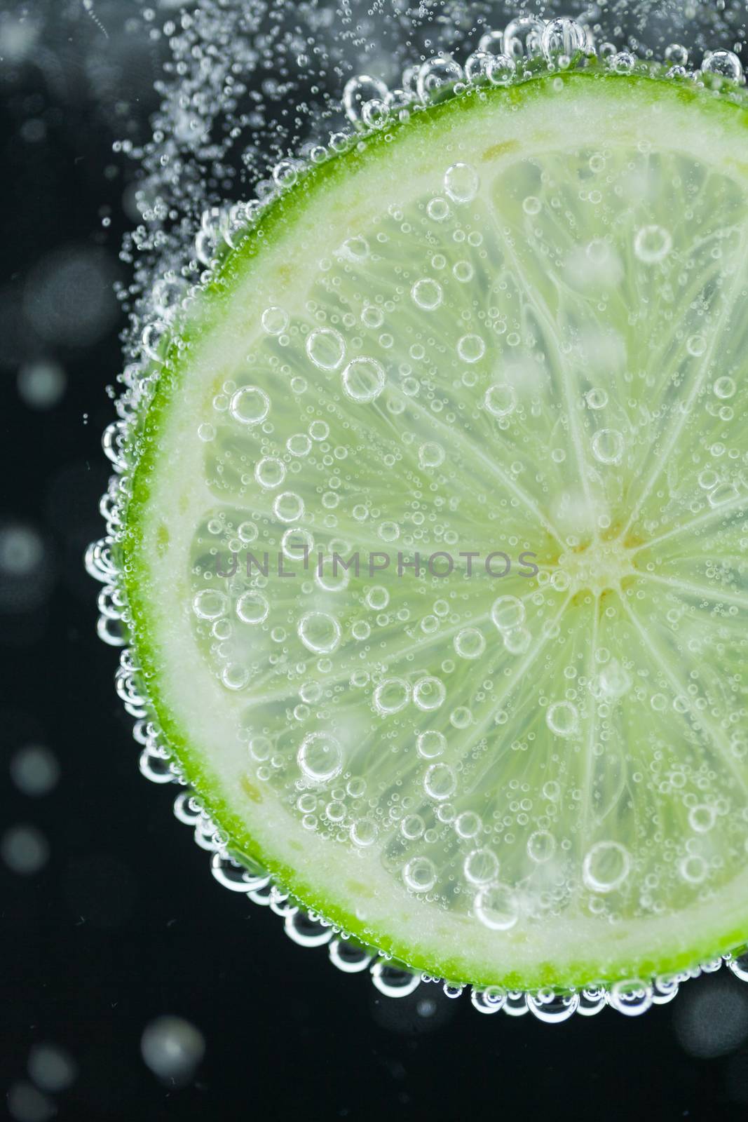 Close-up of single green lime slice falling into carbonated water with bubbles on black background. Refresher drink concept