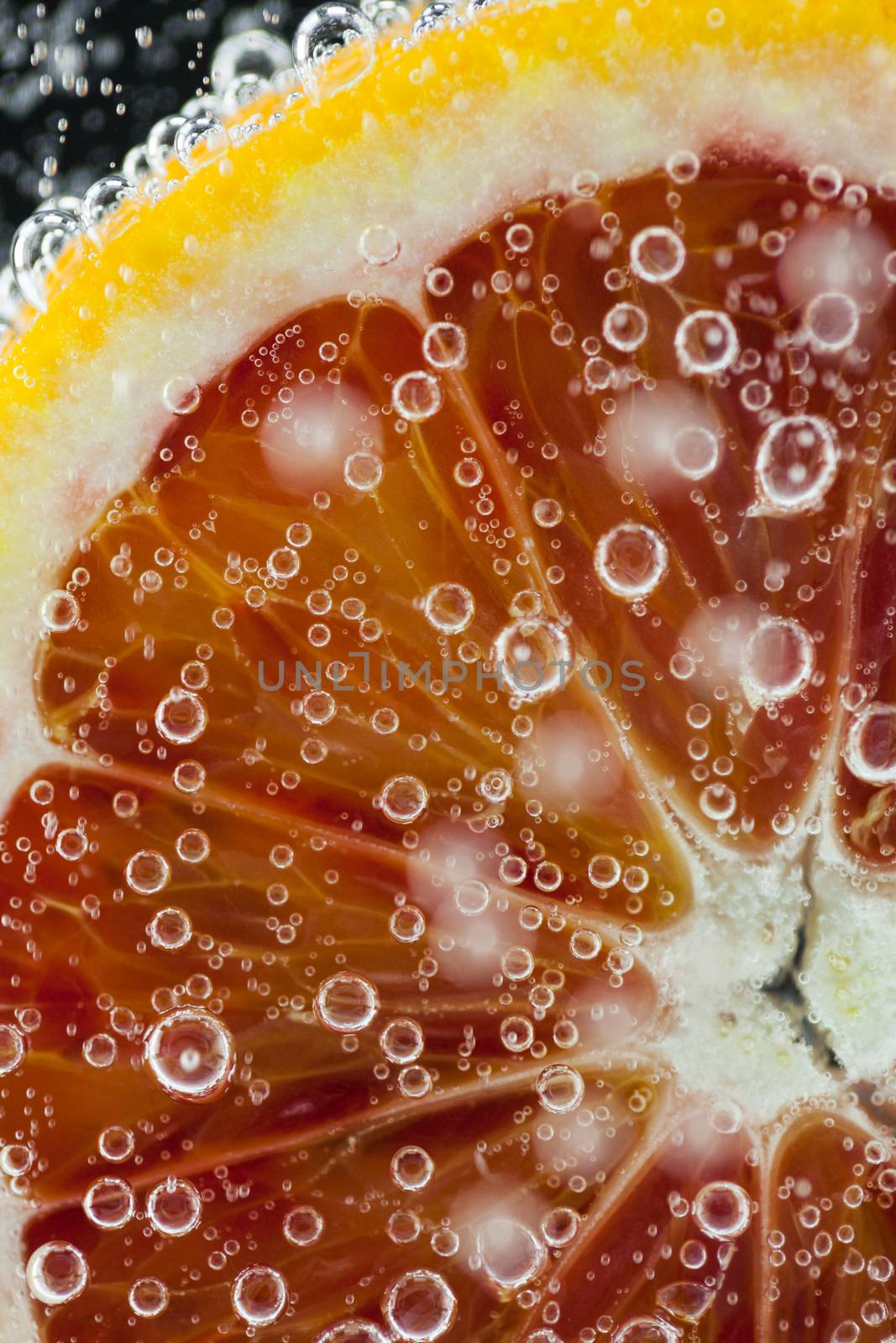 Close-up of blood orange slice diving into carbonated water with bubbles on black background. Refresher drink concept