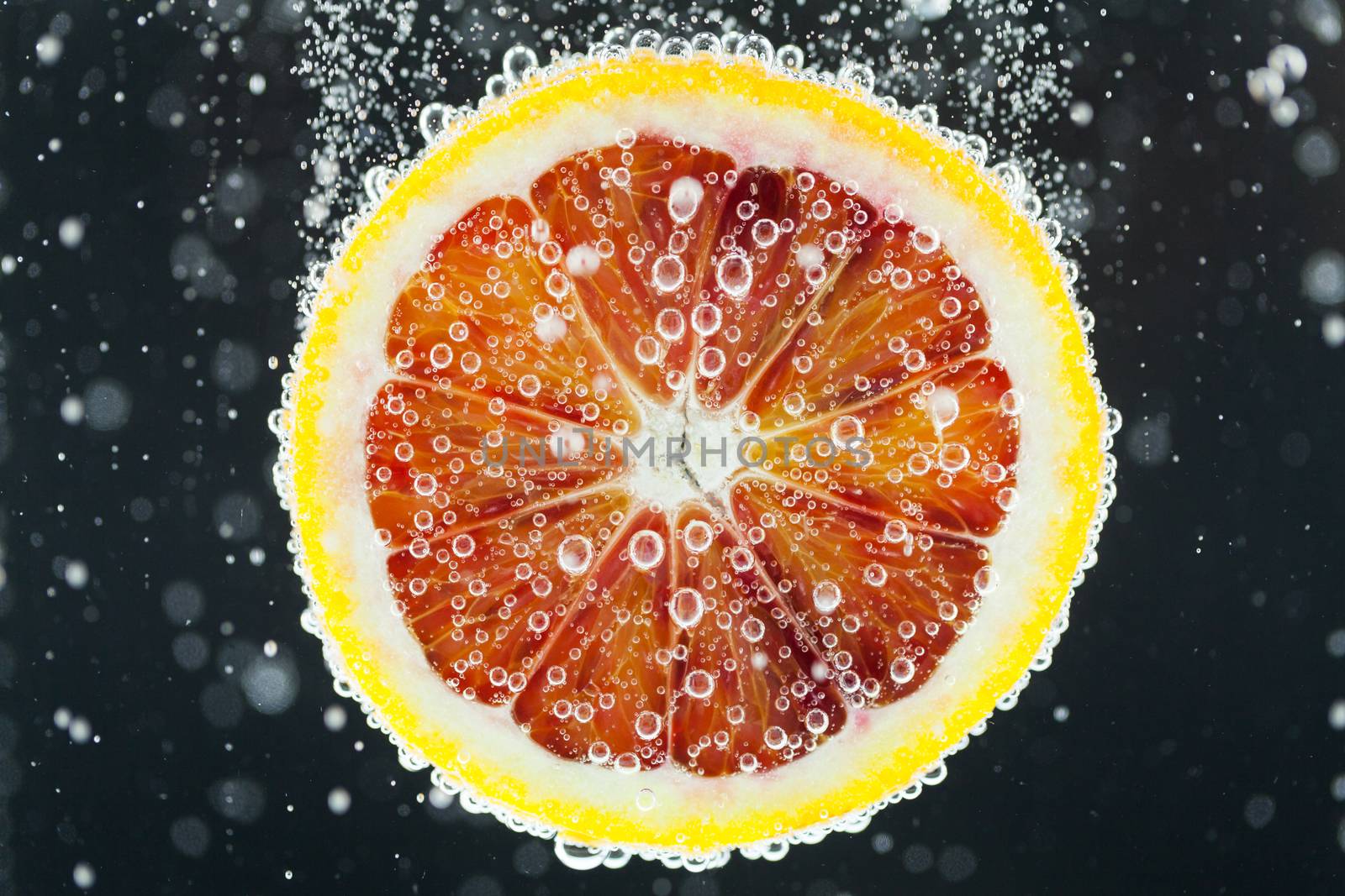 Close-up of blood orange slice diving into carbonated water with bubbles on black background. Refresher drink concept