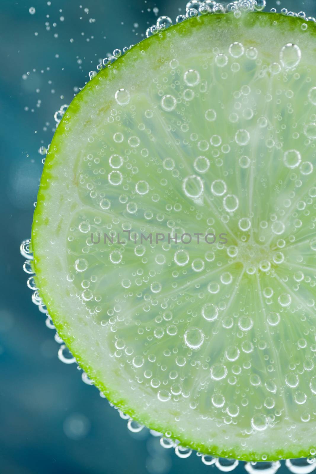 Close-up of lime green slice on blue background diving inside carbonated water with bubbles