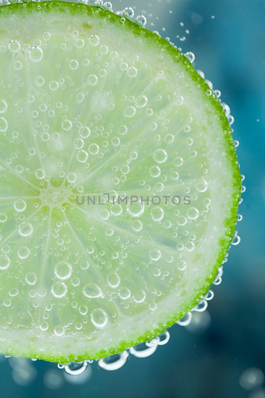 Close-up of lime green slice on blue background diving inside carbonated water with bubbles