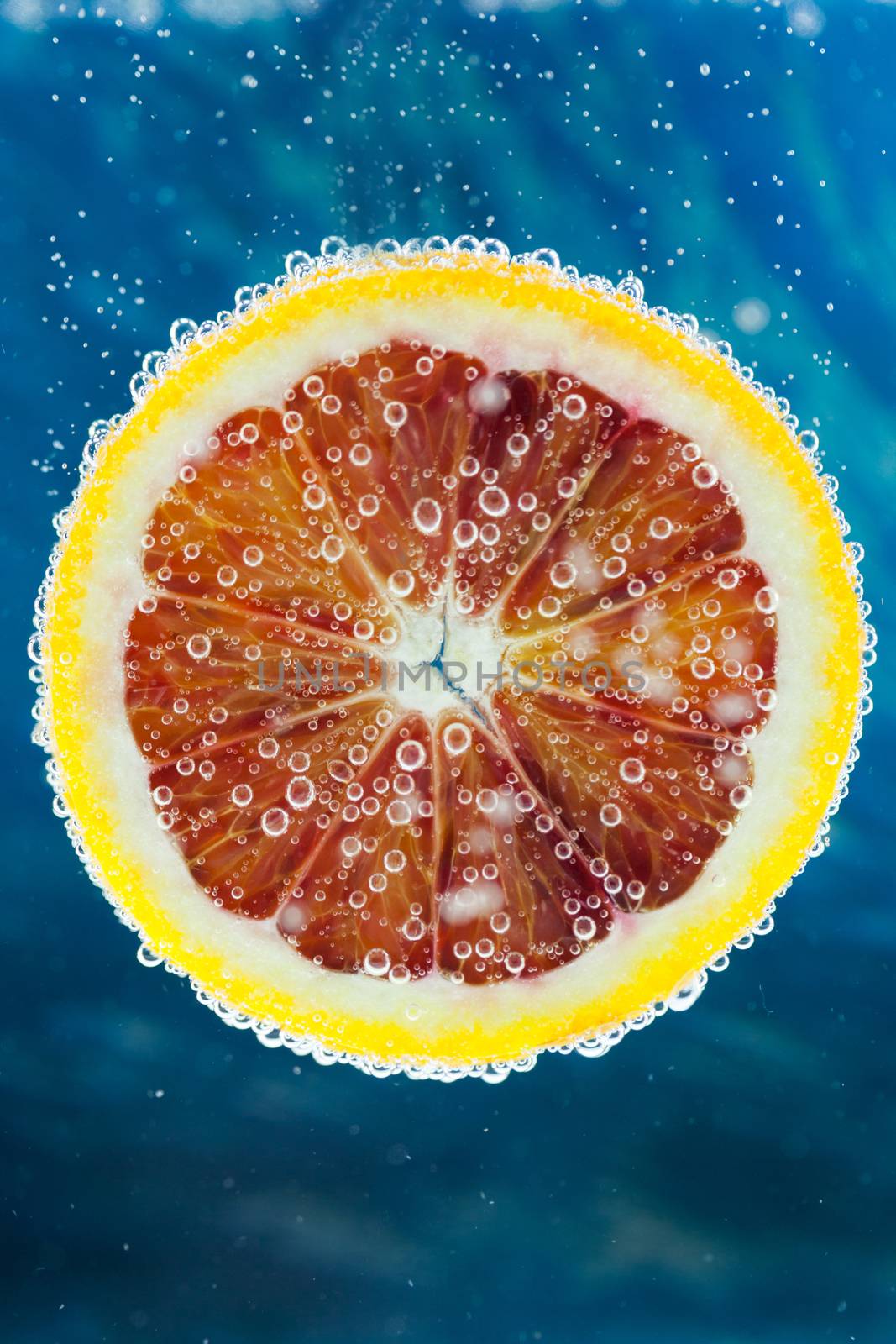 Close-up of slice of citrus blood orange dropped into carbonated water with bubbles on blue background