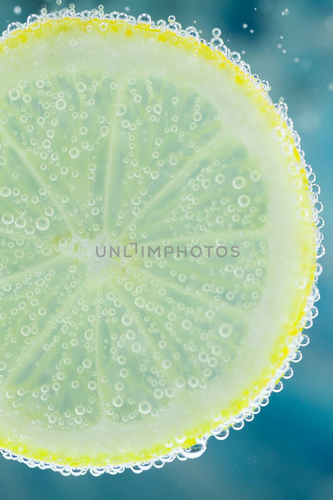 Vertical close-up of slice of lemon falling into carbonated water with bubbles, on blue background. Refresher drink concept