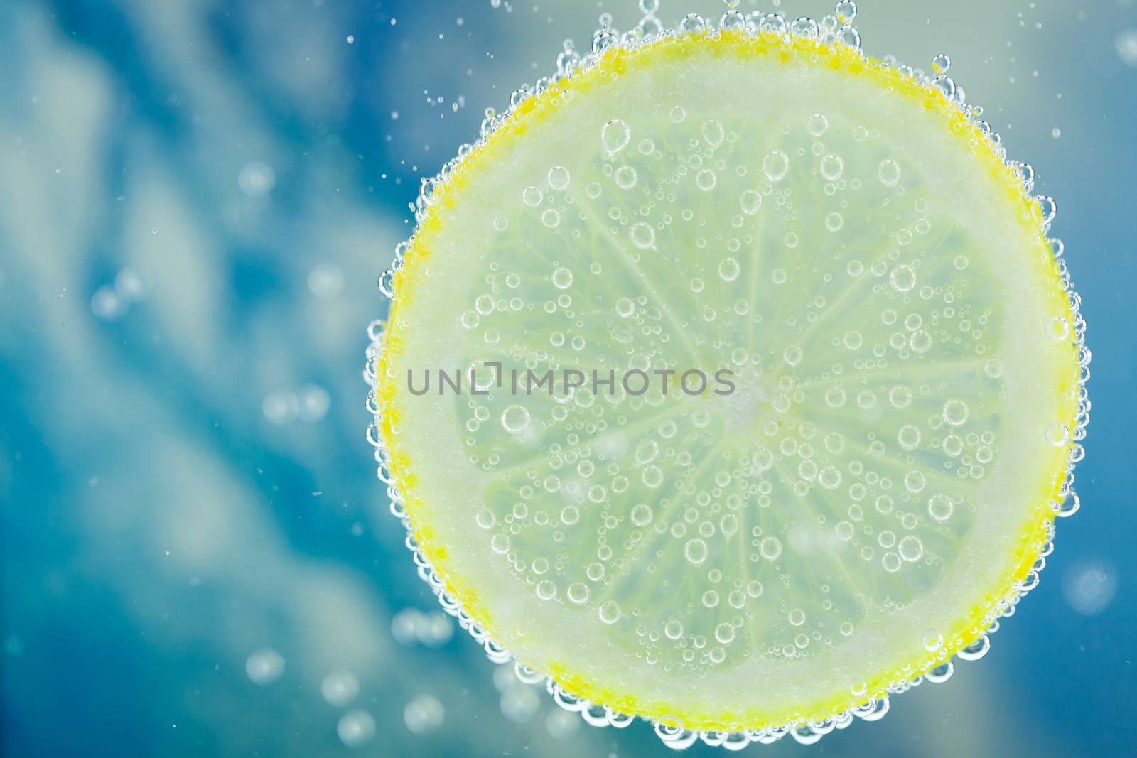 Vertical close-up of slice of lemon falling into carbonated water with bubbles, on blue background. Refresher drink concept