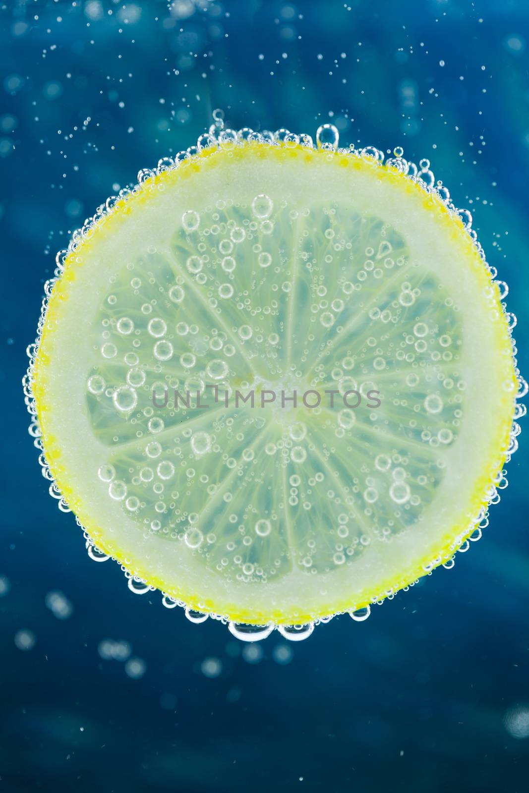 Vertical close-up of slice of lemon falling into carbonated water with bubbles, on blue background. Refresher drink concept