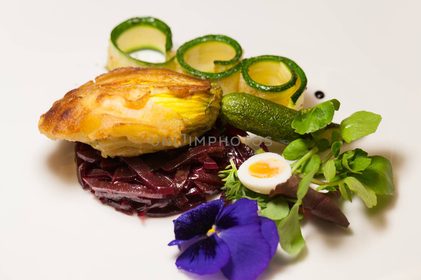 Delicious gourmet plate featuring biscuit sandwich with beets, zucchini strips and pansy garnish over white background