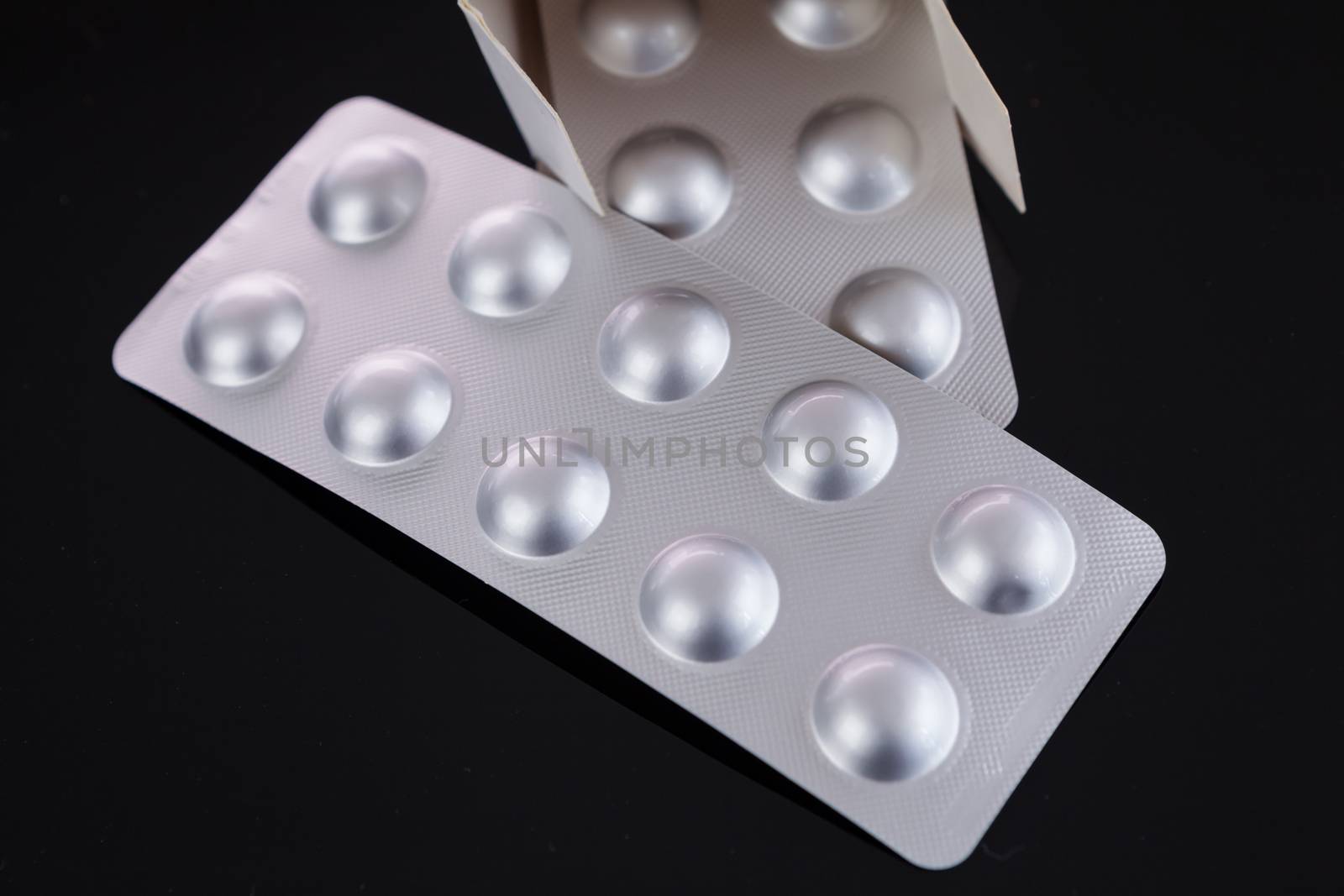 Silver blister pack of small pills together with the a box with additional packs showing through the open lid over a black background in a healthcare concept