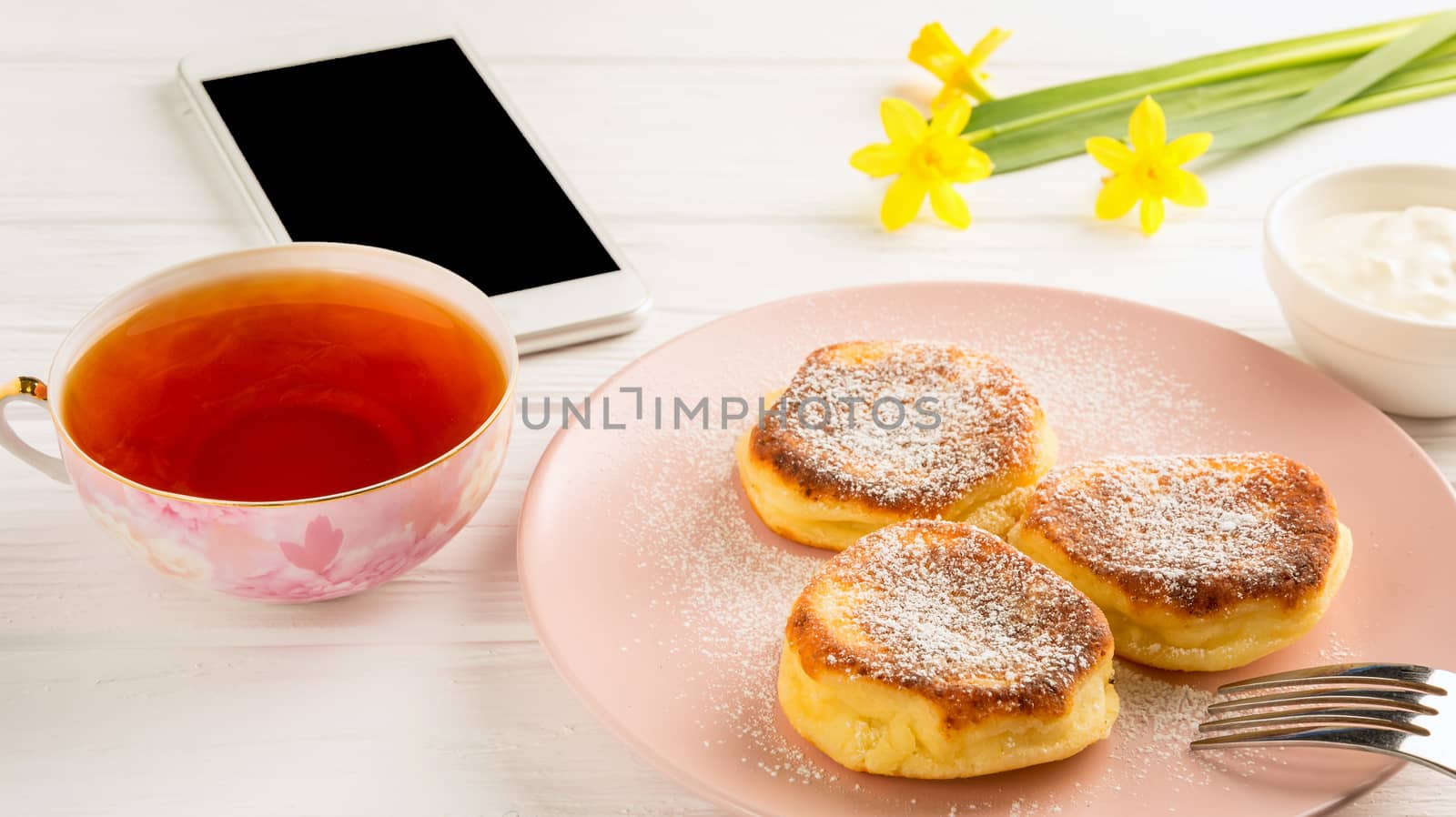 Cheese pancake with powdered sugar, black tea, flowers and smartphone on a white wooden table.