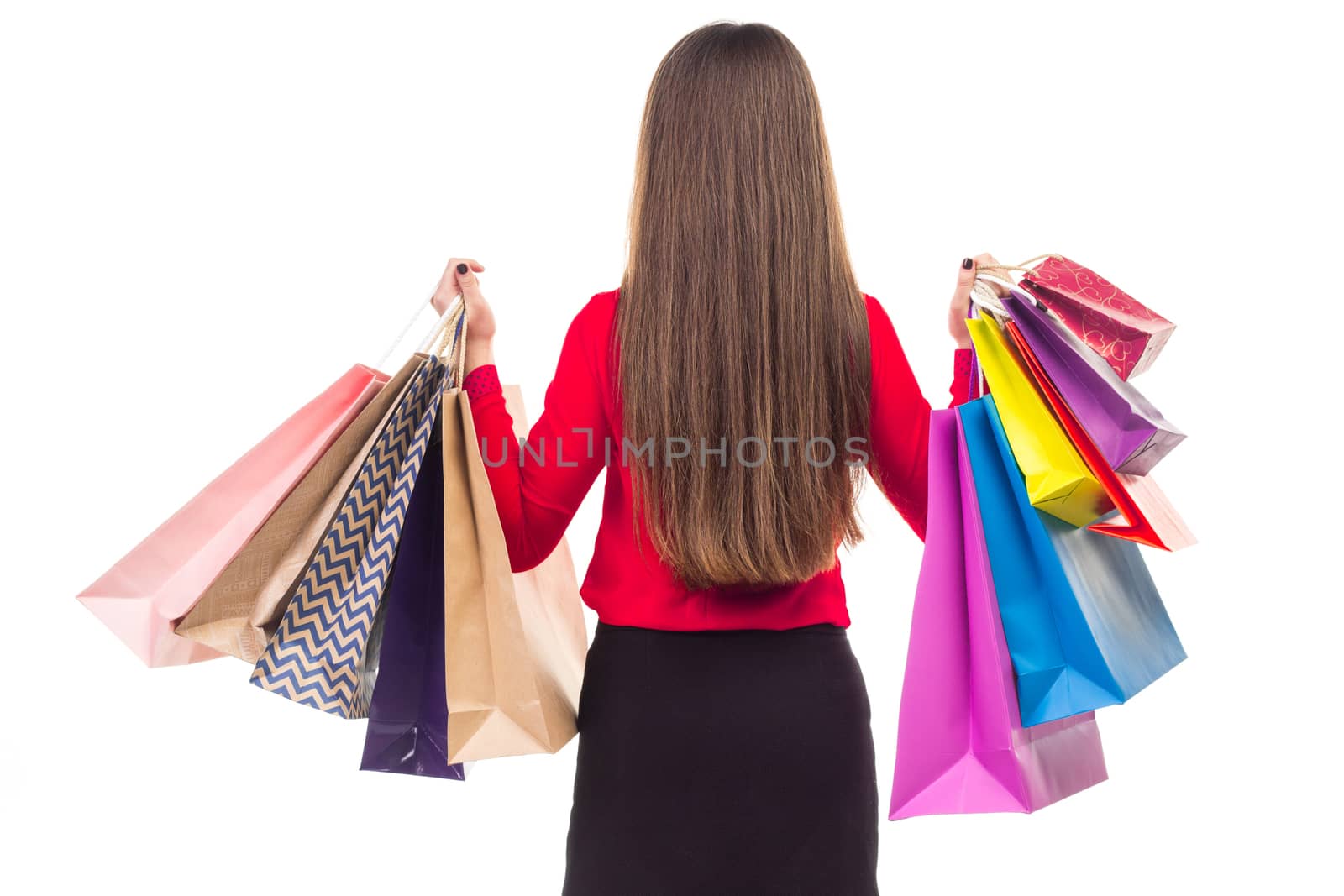 Long-haired young woman wearing red blouse and black skirt from her back holds colourful shopping paper bags