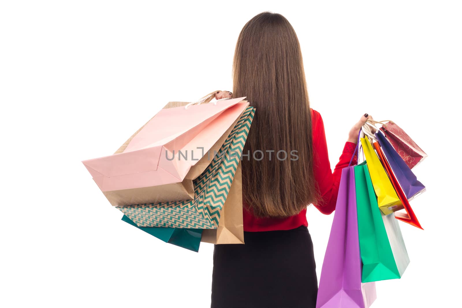 Long-haired young woman wearing red blouse and black skirt from her back holds colourful shopping paper bags