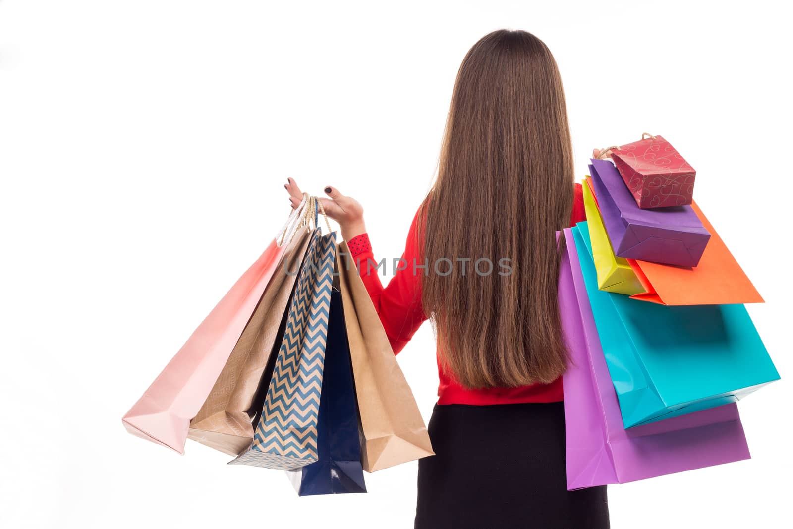 Long-haired young woman wearing red blouse and black skirt from her back holds colourful shopping paper bags
