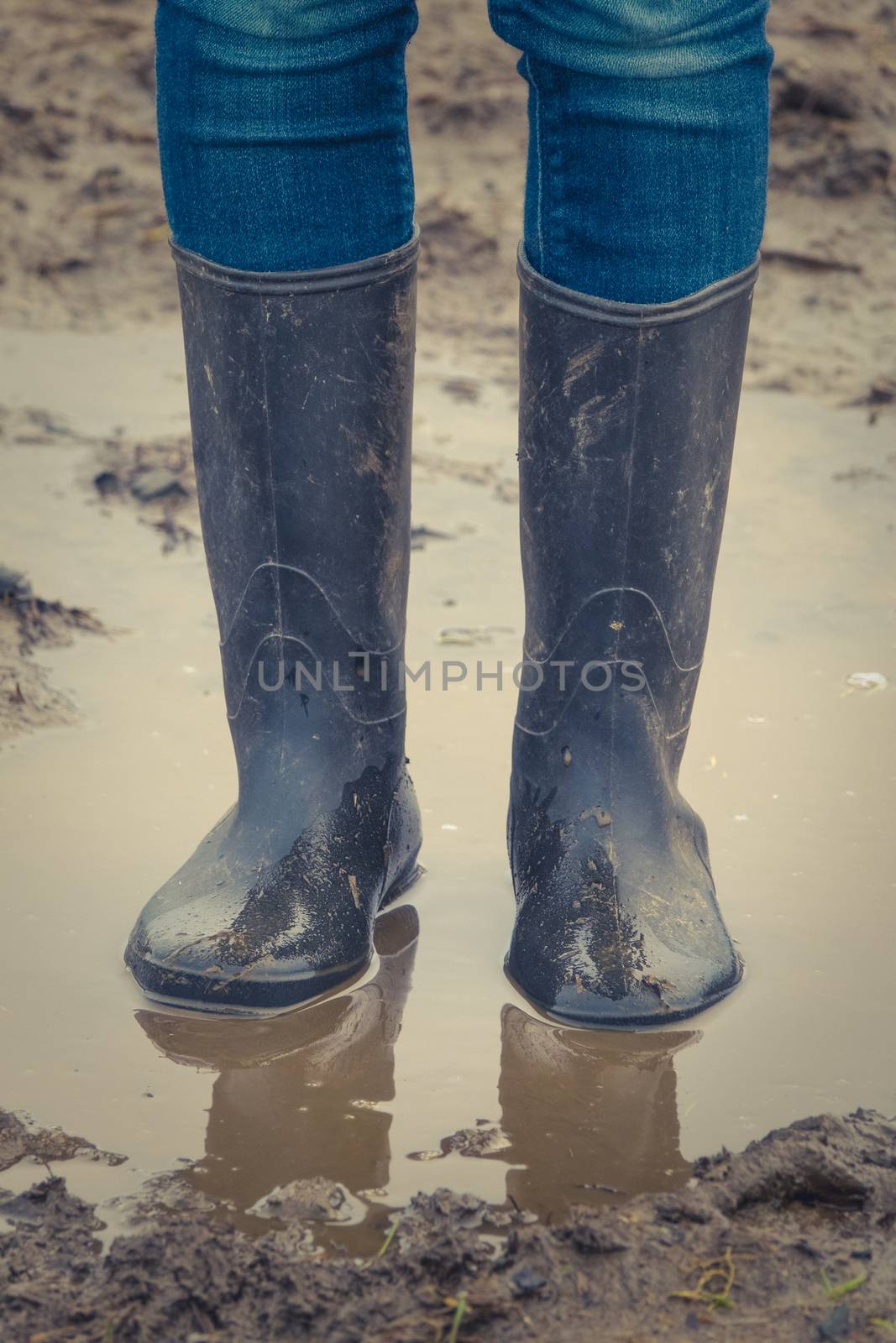 A Child's Legs In Black Rubber Boots Standing In A Muddy Autumn Puddle