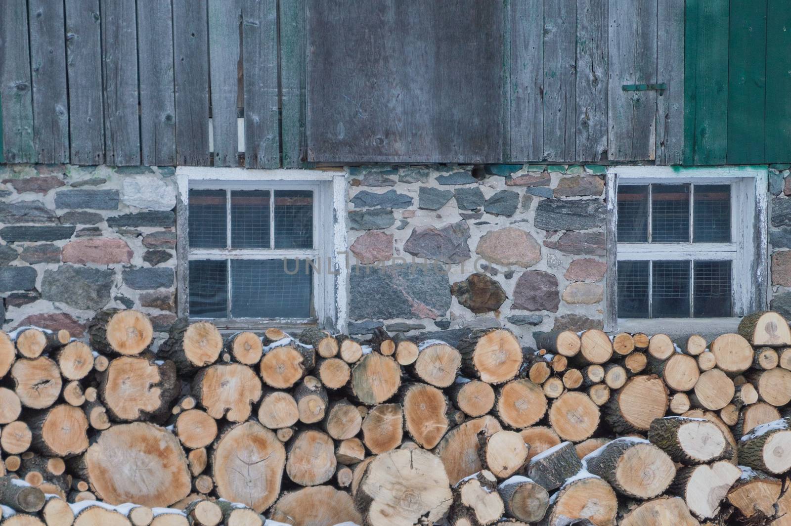 Neatly stacked firewood by an old Ontario Barn.  Barn is barnboards, field stone and has windows too. Snow on the ground.