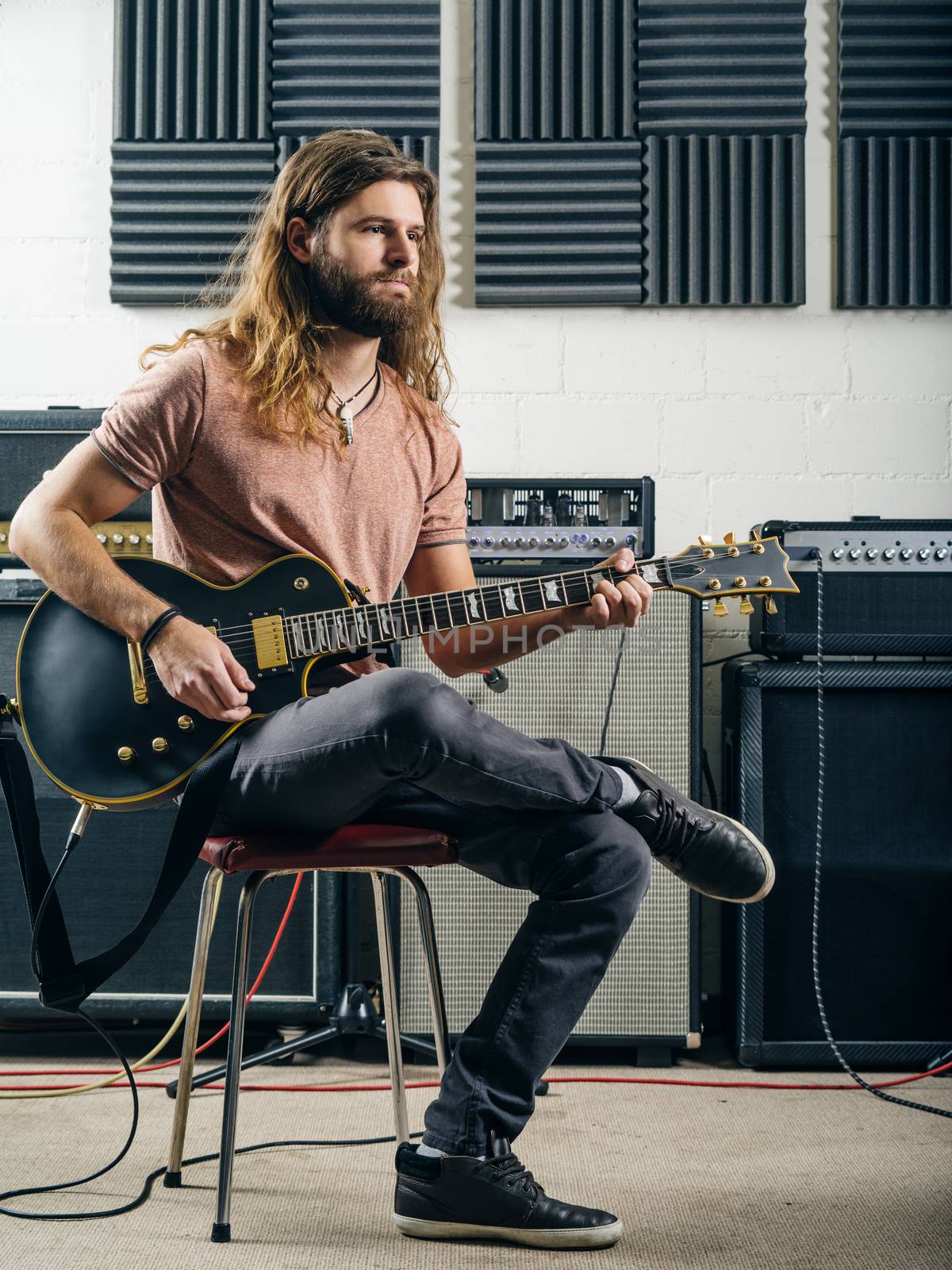 Photo of a young man with long hair and beard playing electric guitar in a recording studio.