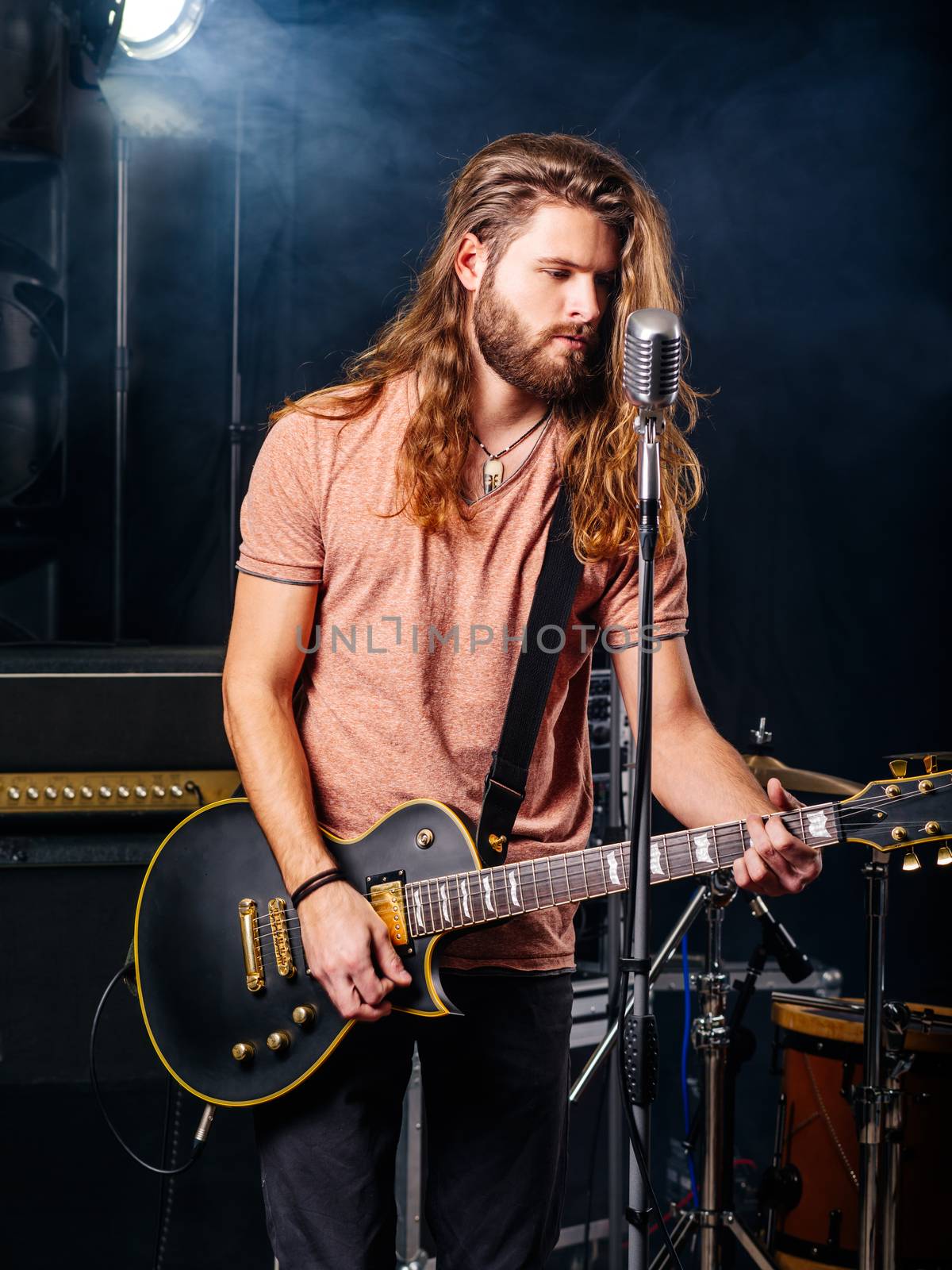 Photo of a young man with long hair and beard playing electric guitar on stage.