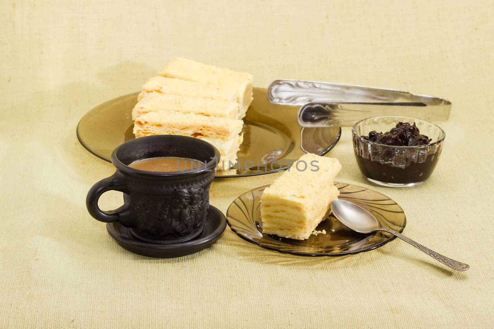 One piece of layered sponge cake on a glass saucer with spoon, several pieces of cake on glass dish, coffee with milk in black cup and jam on a cloth surface
