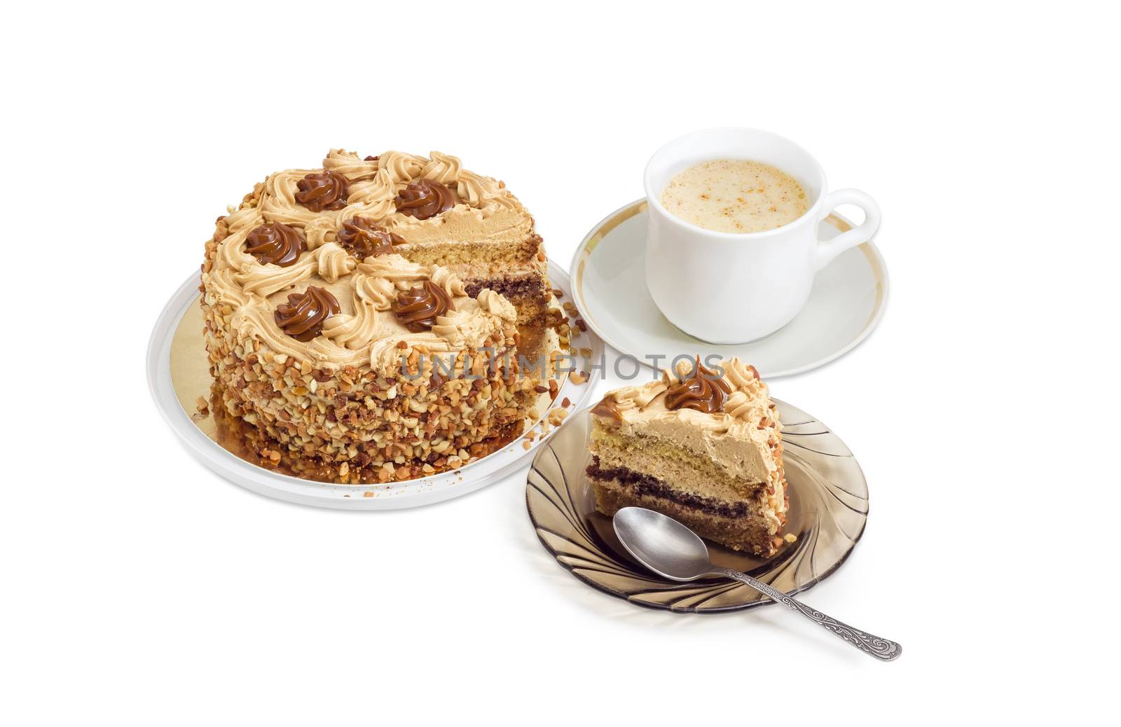 Piece of layered sponge cake, decorated with butter cream, caramelized condensed milk and nuts with spoon on a glass saucer, partly sliced cake and coffee with cream in white cup on a light background
