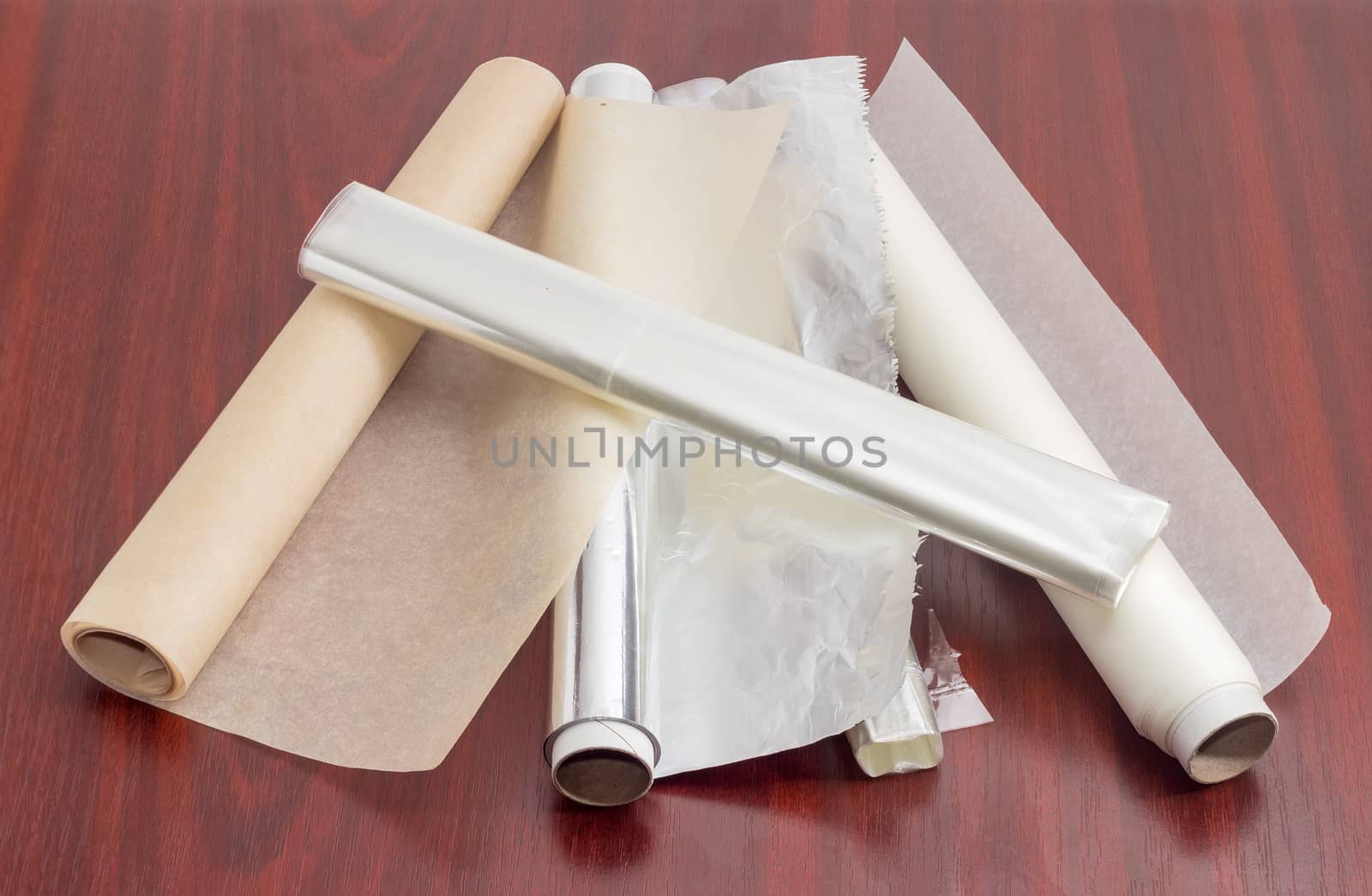 Two rolls of the various parchment paper, two rolls of the plastic oven bags and one roll of the aluminum foil for household use on a dark red wooden surface
