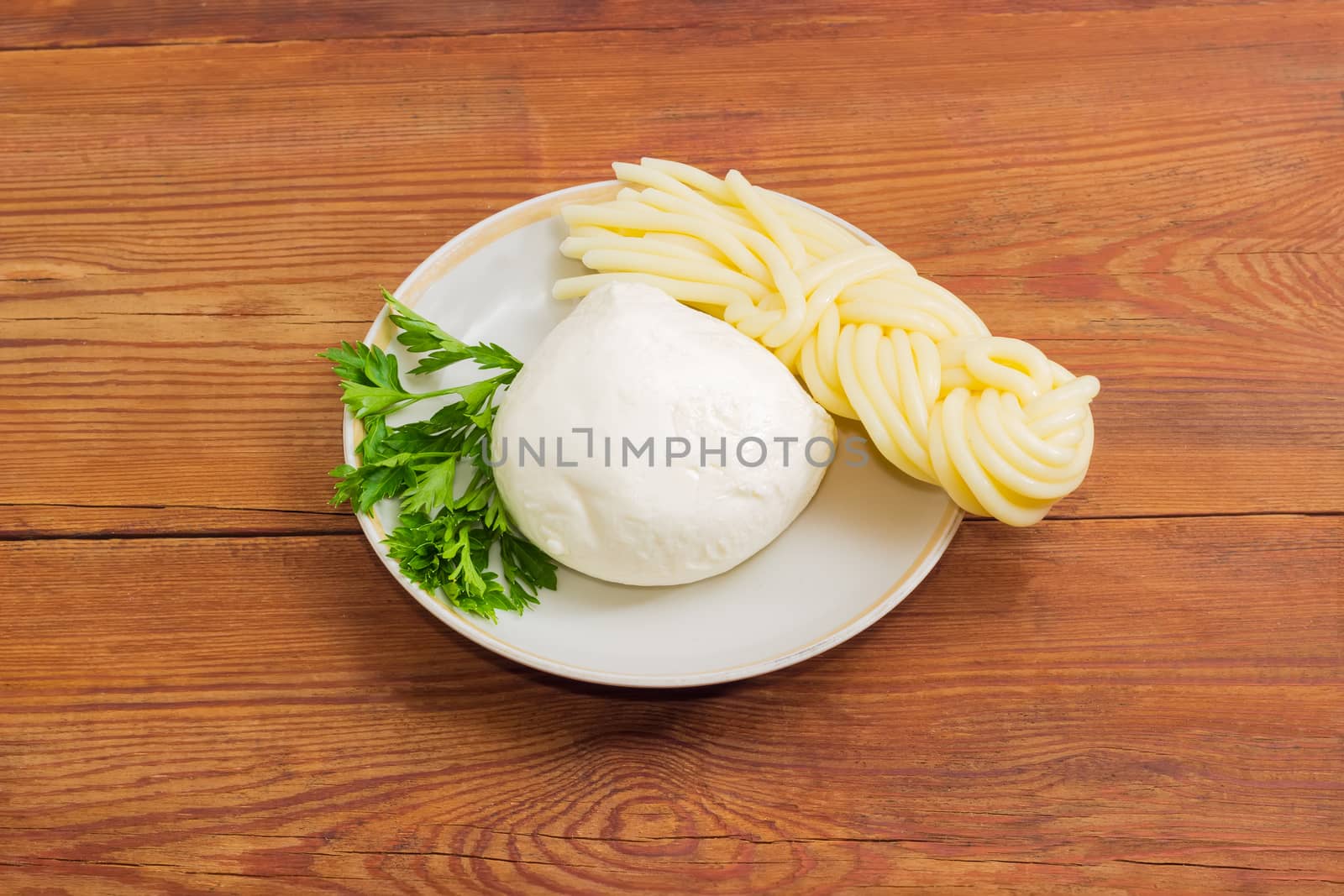 Ball of the fresh mozzarella cheese, portion of the mozzarella cheese in the shape of strings, twisted to form a plait and twig of parsley on the saucer on an old wooden surface
