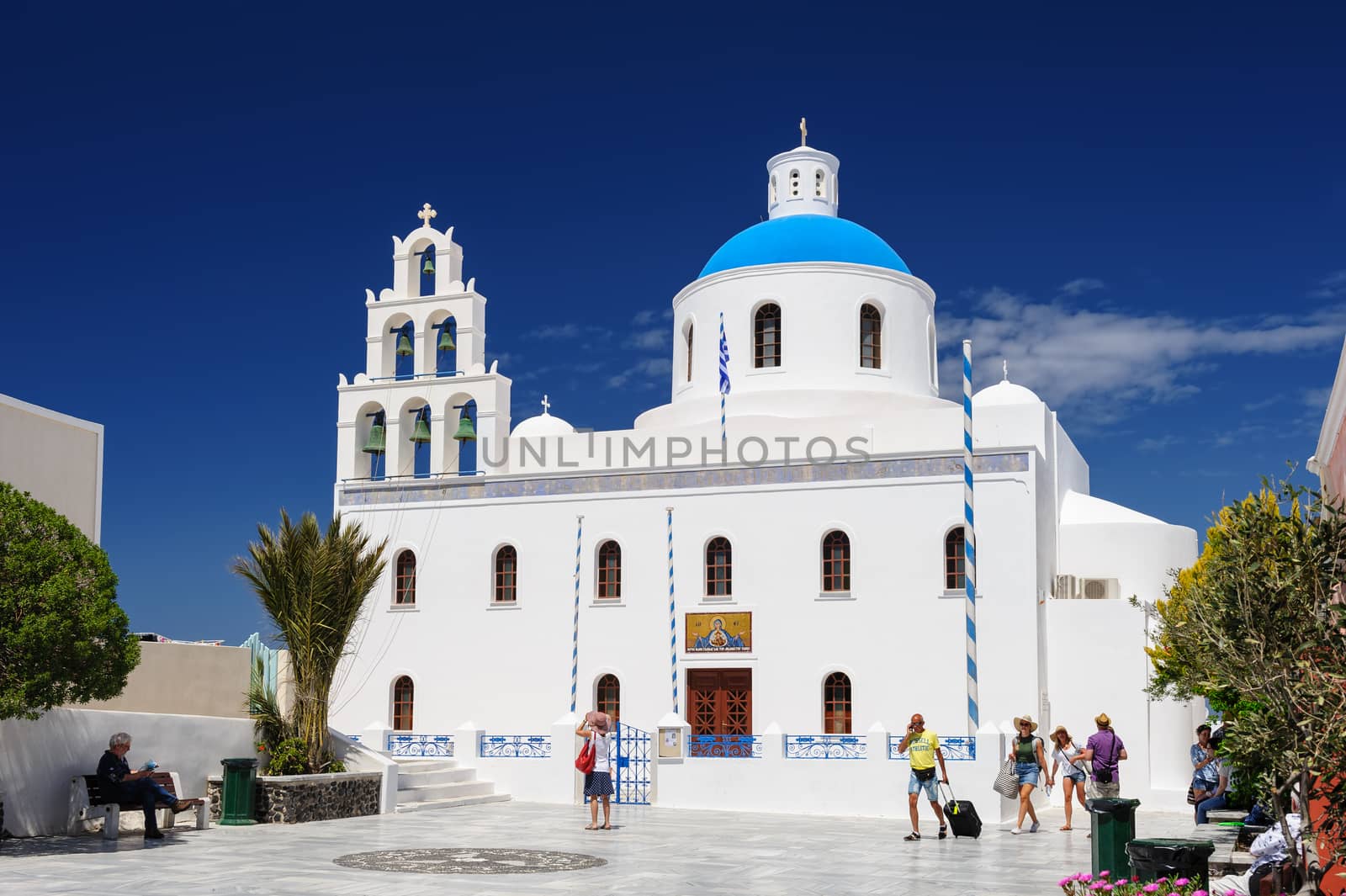 Oia, Santorini island, Cyclades, Greece - April 23, 2016: People relaxing near the Orthodox church of Panagia with bell tower at central square. Oia is a small town and former community in the South Aegean on the islands of Thira AKA Santorini and Therasia, in the Cyclades, Greece.