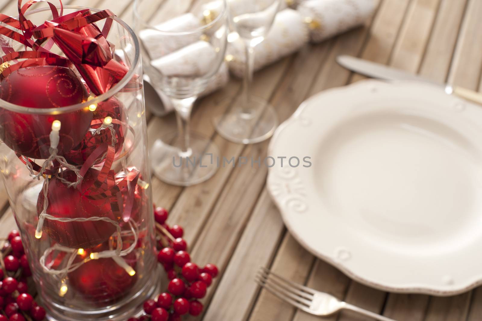 Christmas place setting with red themed decorations in a glass container surrounded by colorful red berries with a white empty plate, glassware, utensils and crackers