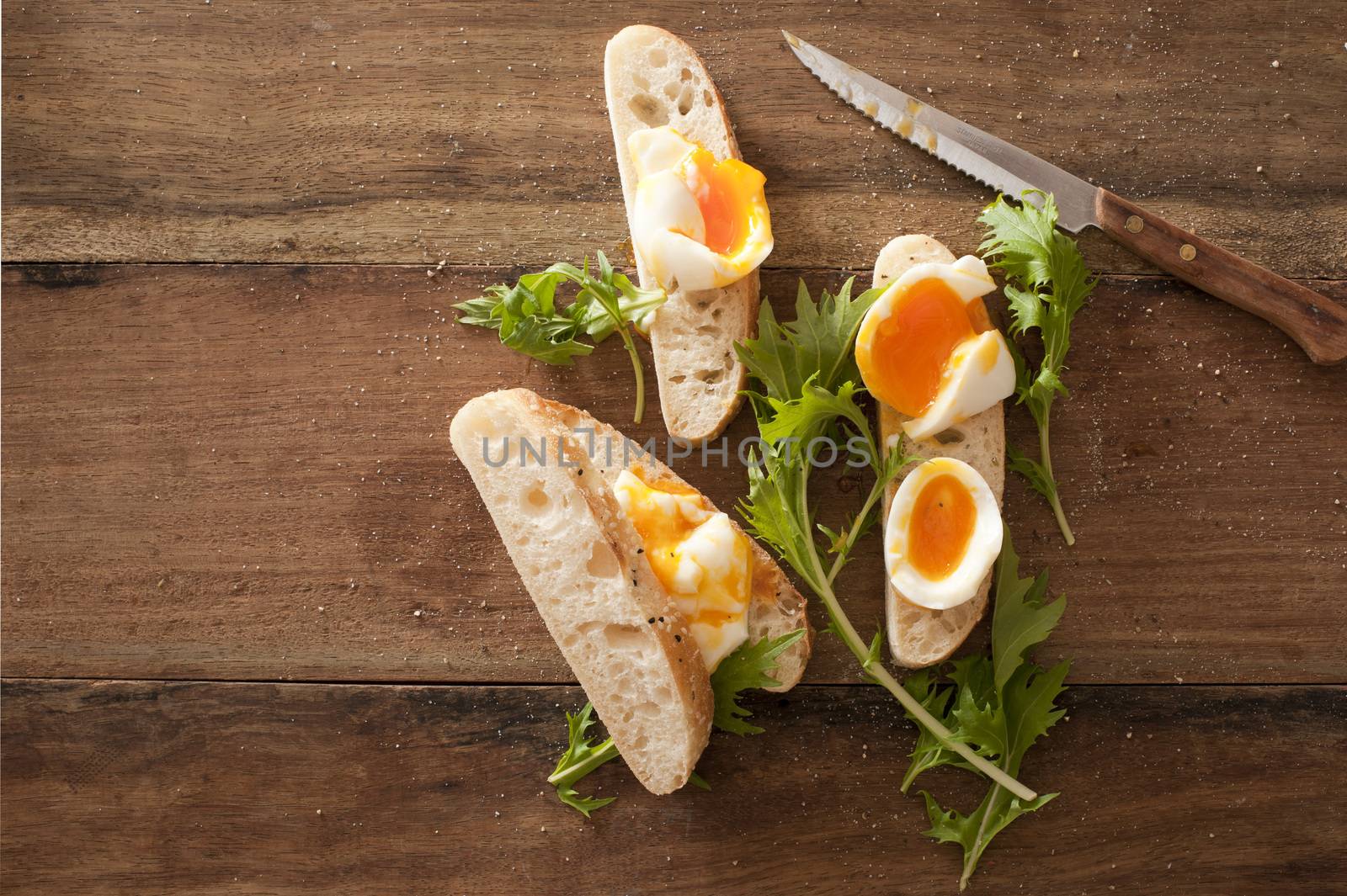 Sliced soft boiled eggs on freshly baked baguette with fresh green coriander and a knife on a wooden kitchen table