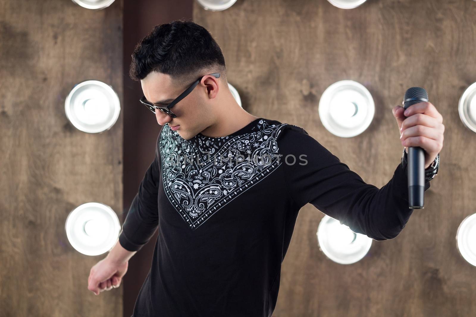 Male singer of rock or pop music dressed in black and sunglasses with microphone performs on scene with lightening projectors on background