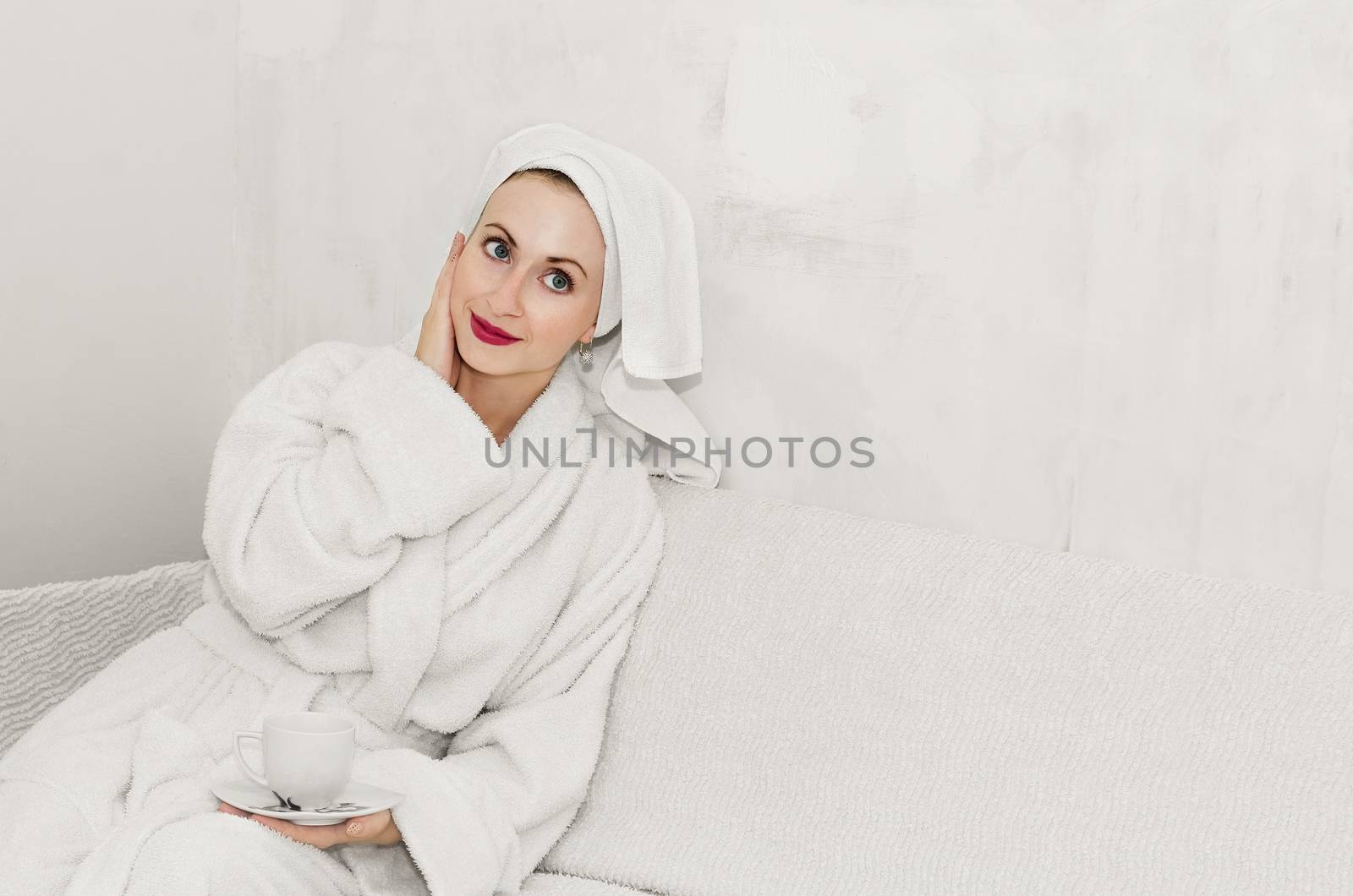 Beautiful girl sitting on the sofa in a fluffy Bathrobe in a room with no Wallpaper on the walls. White background, high key. by Gaina