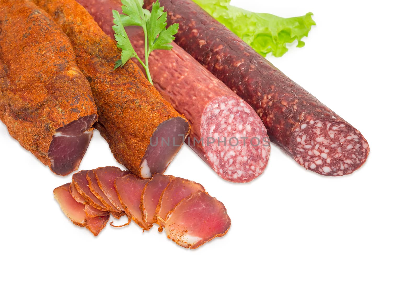 Fragment of the partly sliced two pieces of the dried pork tenderloin and two different dry smoked sausage with parsley twig on the lettuce closeup on a light background  
